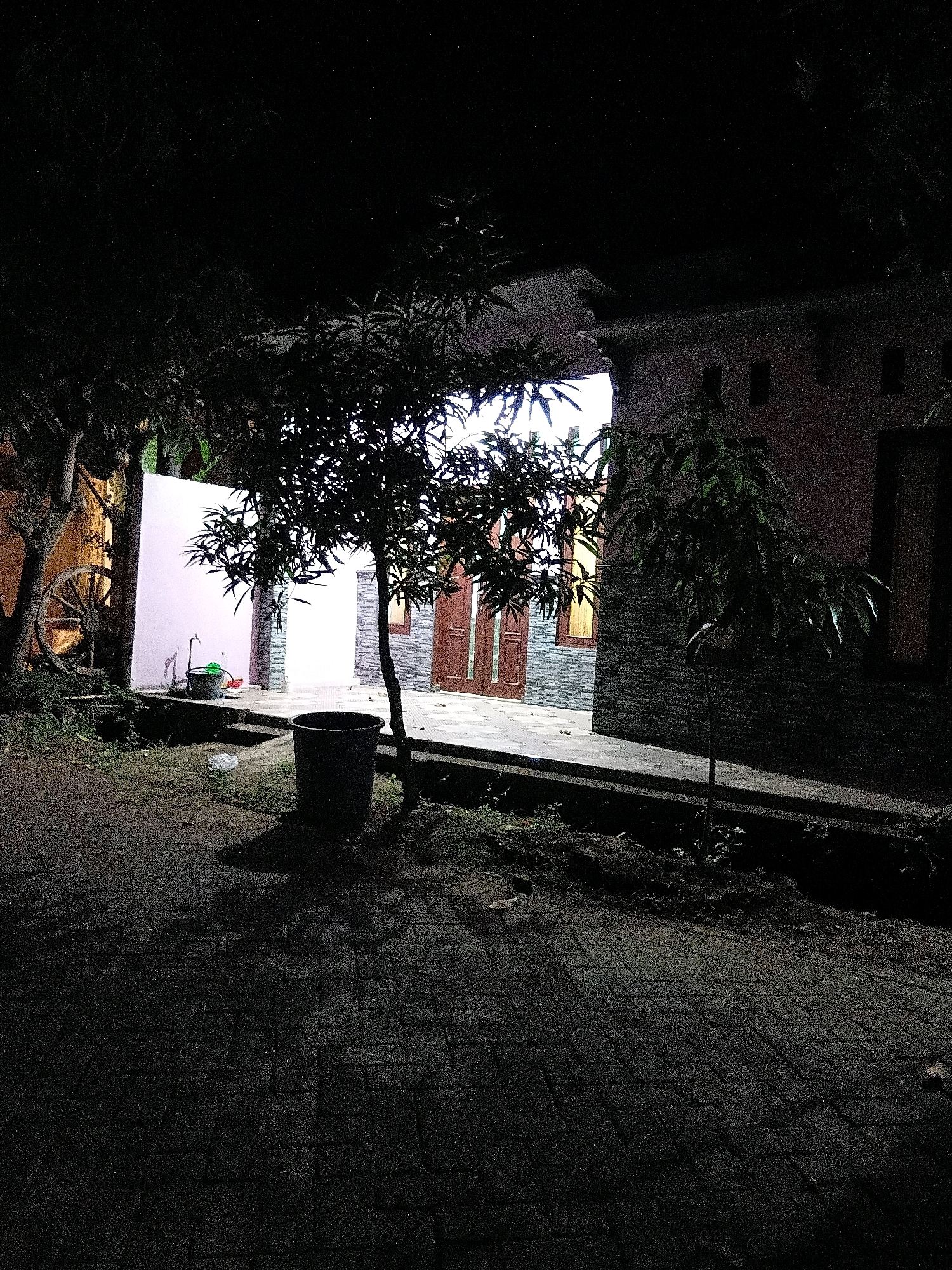 The image depicts an outdoor scene at night, likely in a residential area of Cikal, Indonesia. In the foreground, a paved pathway made of interlocking bricks runs diagonally from the bottom left to the upper right. To the left of the pathway, there is a patch of dark ground with some foliage.
Dominating the mid-ground is a dark-colored trash bin. Behind it, a small tree with sparse leaves casts a shadow on the pathway. Further back, a white wall is visible, with a large wooden wagon wheel leaning against it on the far left. Near the wagon wheel, there is a dark-colored bucket with a handle and a spout, next to a tap or faucet.
To the right of the white wall, a building is partially visible. The building has a reddish-brown facade with horizontal bands of stone or brickwork. A dark-colored door with a wooden frame is visible, and to the right of the door, a window with a wooden frame and visible panes of glass. Another, smaller tree with dark leaves stands in front of the building on the right side. The overall lighting suggests it is nighttime, with artificial light sources illuminating parts of the scene, creating deep shadows. The environment appears to be a yard or courtyard adjacent to a residential structure.