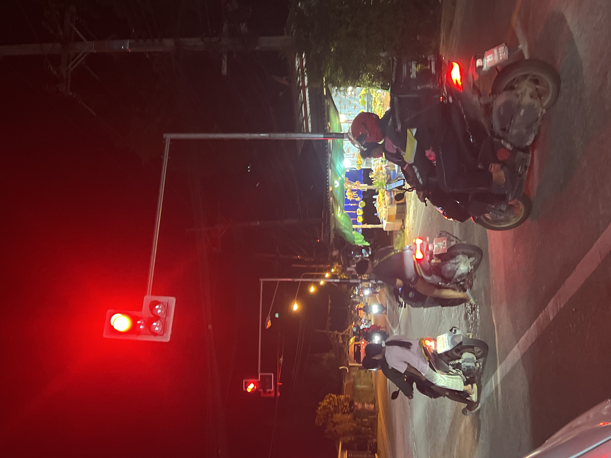 The image depicts a street scene at night in Ko Pha-ngan District, Thailand, illuminated by a red traffic light. Several motorbikes are on the road, with people riding them. In the foreground, a motorbike carries two people, the passenger with long braided hair wearing a light-colored shirt and striped pants. Another motorbike behind it has a person wearing a red helmet and a dark jacket with yellow accents, equipped with a large black delivery box on the rear. Other motorbikes are visible further down the road, some with illuminated headlights. The background shows buildings and stalls with bright lights, likely businesses, and a utility pole with wires. The overall atmosphere is that of a busy nocturnal street.