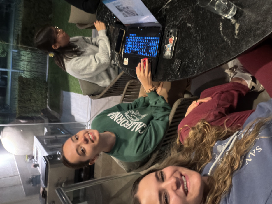 The scene depicts three individuals gathered around a dark, polished table within an indoor-outdoor living space, likely in San Bernardino Tlaxcalancingo, Mexico, during evening hours. In the foreground on the left, a woman with long, wavy brown hair, wearing a light blue hooded sweatshirt, maroon pants, and white sneakers with black accents, smiles towards the camera. Adjacent to her, a second woman with dark hair pulled back wears a green sweatshirt with "CALIFORNIA" text and tan pants; she is also smiling at the camera while her right hand, adorned with a light-colored bracelet, operates a red computer mouse connected to an open laptop. The laptop displays a glowing blue keyboard, text on its left screen portion, and an image or document on the right. On the opposite side of the table, a third woman with dark hair and glasses, wearing a light grey sweater, is observed looking downwards, focused on the table. A clear phone case with a colorful sticker, a dark remote control, and a clear drinking glass are also present on the table. The background reveals an outdoor kitchen area with a sink and counter to the left, while beyond a clear glass partition, a manicured lawn, hedge rows, and a dark fence are visible.