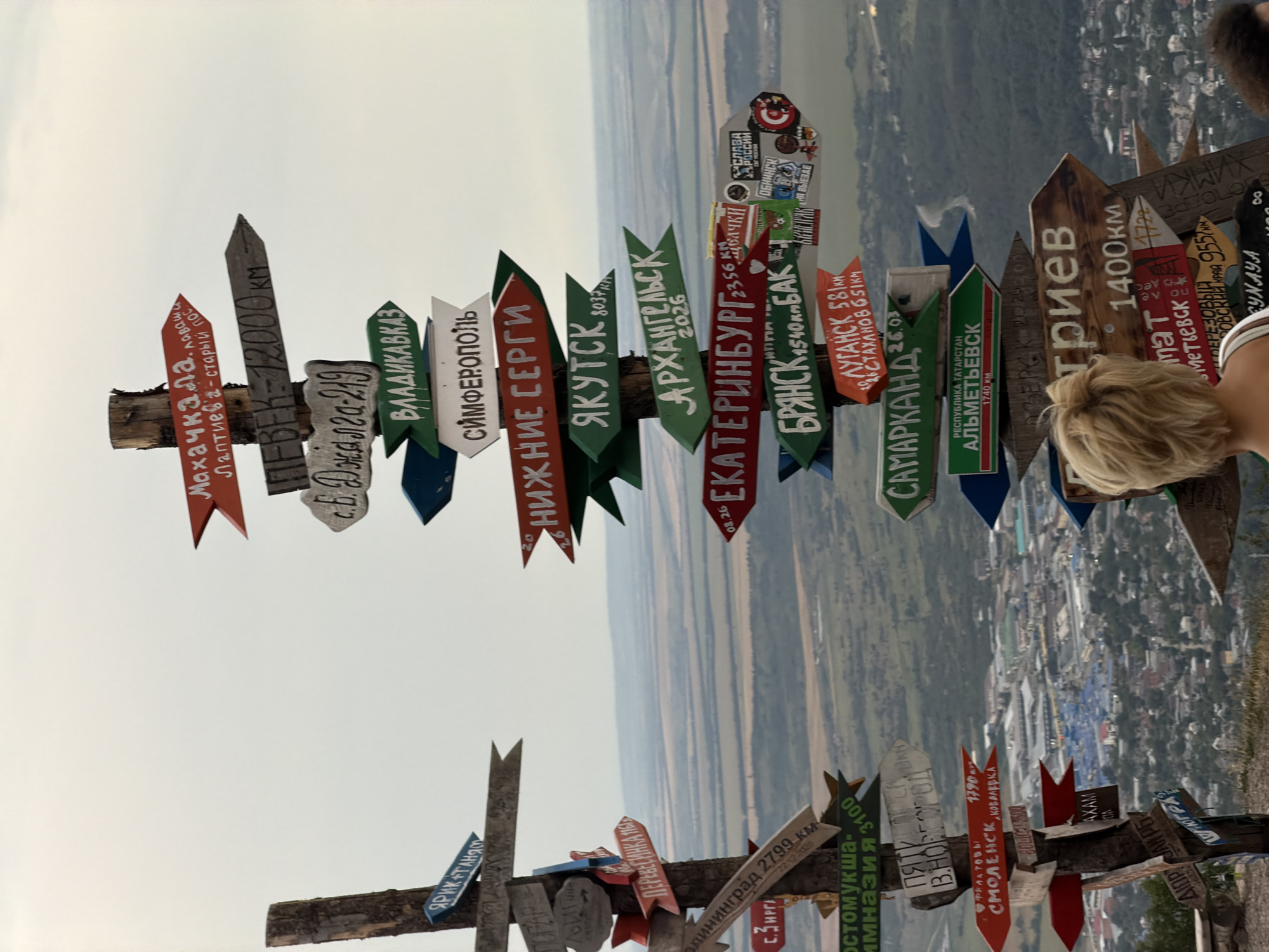 This is a medium shot, eye-level, outdoor photograph taken on an overcast day. The main subject is a weathered wooden signpost densely populated with numerous colorful directional signs pointing in various directions. These signs are made of wood and painted with white and red lettering, indicating destinations and distances in kilometers, many of which are in Russian. Examples of visible destinations include "Махачкала," "Лермонтов," "Пятигорск," "Самарканд," "Екатеринбург," "Брянск," "Якутск," and "Симферополь." The signpost is positioned on a rocky outcropping overlooking a vast, hazy valley with a city visible in the distance. The city features a mix of residential and industrial buildings, with roads winding through the landscape. Two individuals are partially visible in the foreground, with their backs to the camera. The woman on the left has short, blonde hair and is wearing a white top. The person on the right has dark, curly hair. The image appears to capture a scenic overlook in the vicinity of the city of Khoroshovskiy, Russia, known for its picturesque landscapes.