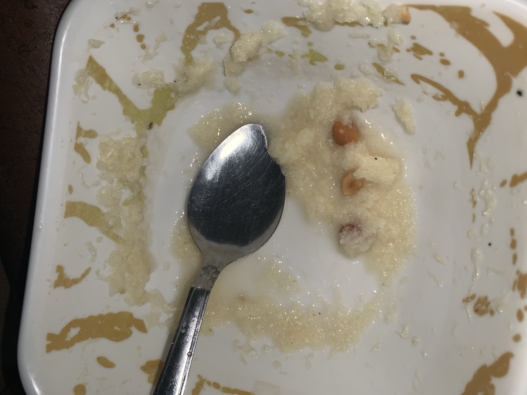 The image presents an overhead view of a white, square-shaped plate with rounded corners, containing the remnants of a recently consumed meal. A silver spoon rests diagonally across the plate, its bowl positioned within a cluster of the leftover food.
The food remnants consist of a light-colored, granular or semi-liquid substance, resembling a porridge or gruel. Visible within these remnants are several whole and broken pieces of what appear to be peanuts or similar legumes. Small, dark specks are scattered across the food and the plate's surface, possibly indicating spices or minor debris.
The plate itself bears smears and streaks of a yellowish-brown substance, likely a sauce or oil, along its edges and across the bottom, suggesting the plate was used for a saucy or oily dish. The overall scene depicts a plate immediately after a meal, with food residue and the spoon still present before cleaning.
Given the location context of Rafin Par, Nigeria, the dish could be a local delicacy such as 'kunun gyada' (peanut porridge) or another grain-based meal common in the region. No individuals are visible in the image, nor is there any discernible text. The time of day and external weather conditions cannot be determined from the photograph. The background surrounding the plate is dark and indistinct, possibly a wooden table or countertop.