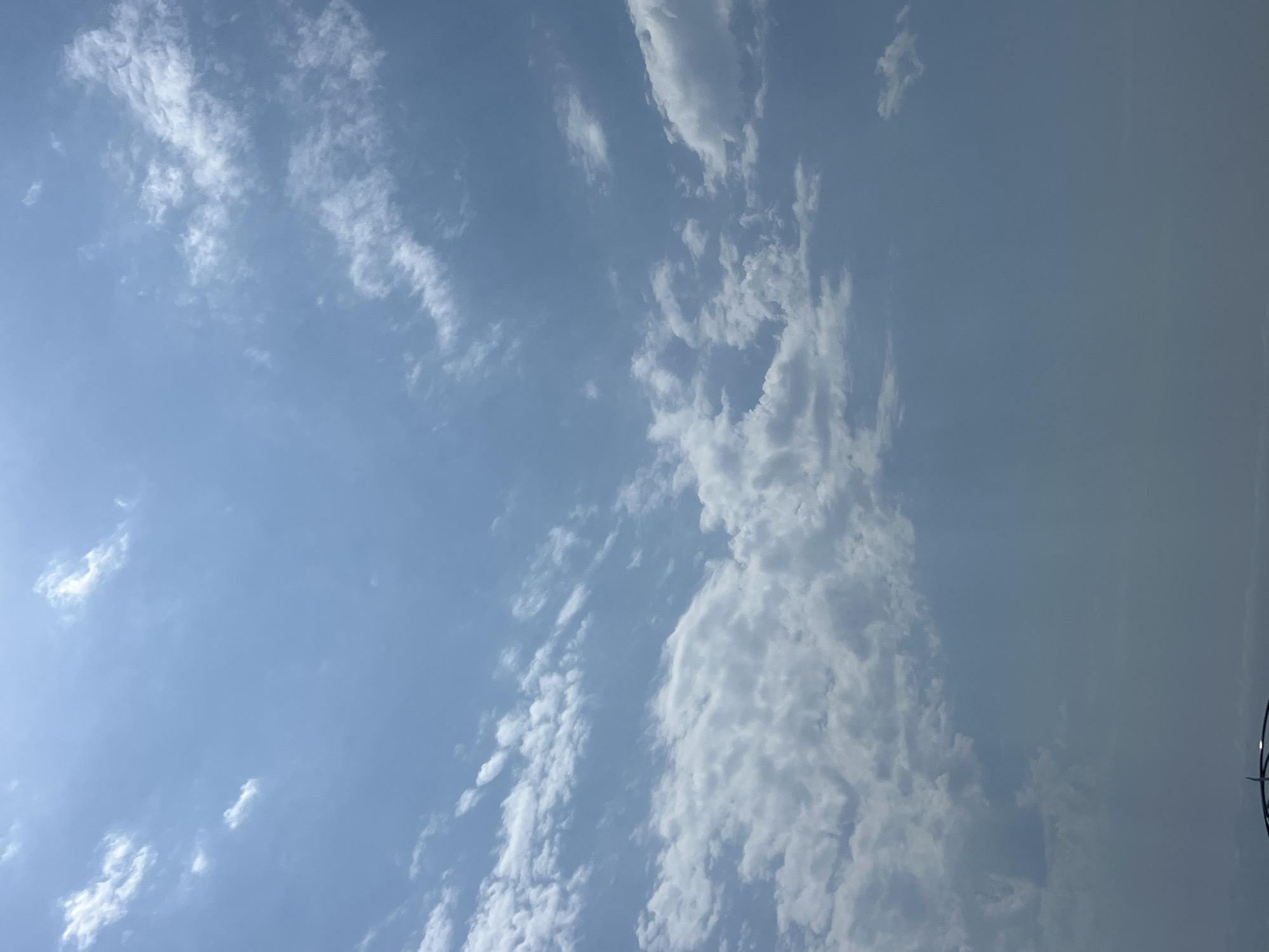 The image shows a sky with scattered white clouds against a light blue background. The sun appears to be behind the clouds, creating a bright, hazy effect. There is a hint of a structure, possibly a building or an elevated walkway, visible at the very bottom of the frame, with a dark curved element that could be part of a railing or a roof. The scene is likely an outdoor environment, potentially a park, viewpoint, or a coastal area, given the presence of a structure at the edge of the frame and the expansive sky. Without further information or context, it is not possible to definitively identify the location as Lermontovo, Russia.