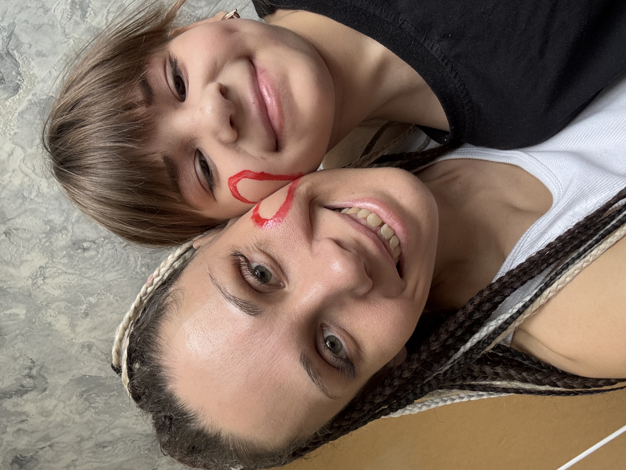 The image depicts two smiling individuals, likely a mother and child, lying close together. The child, in a black shirt, has short, straight brown hair with bangs. The adult, wearing a white ribbed tank top, has braided hair with a mix of brown and blonde strands. Both have a red heart shape drawn on their cheek. The background appears to be a gray textured surface on one side and a plain brown surface on the other. The image does not provide enough information to confirm its location in Rusanovo, Russia, or any specific city.