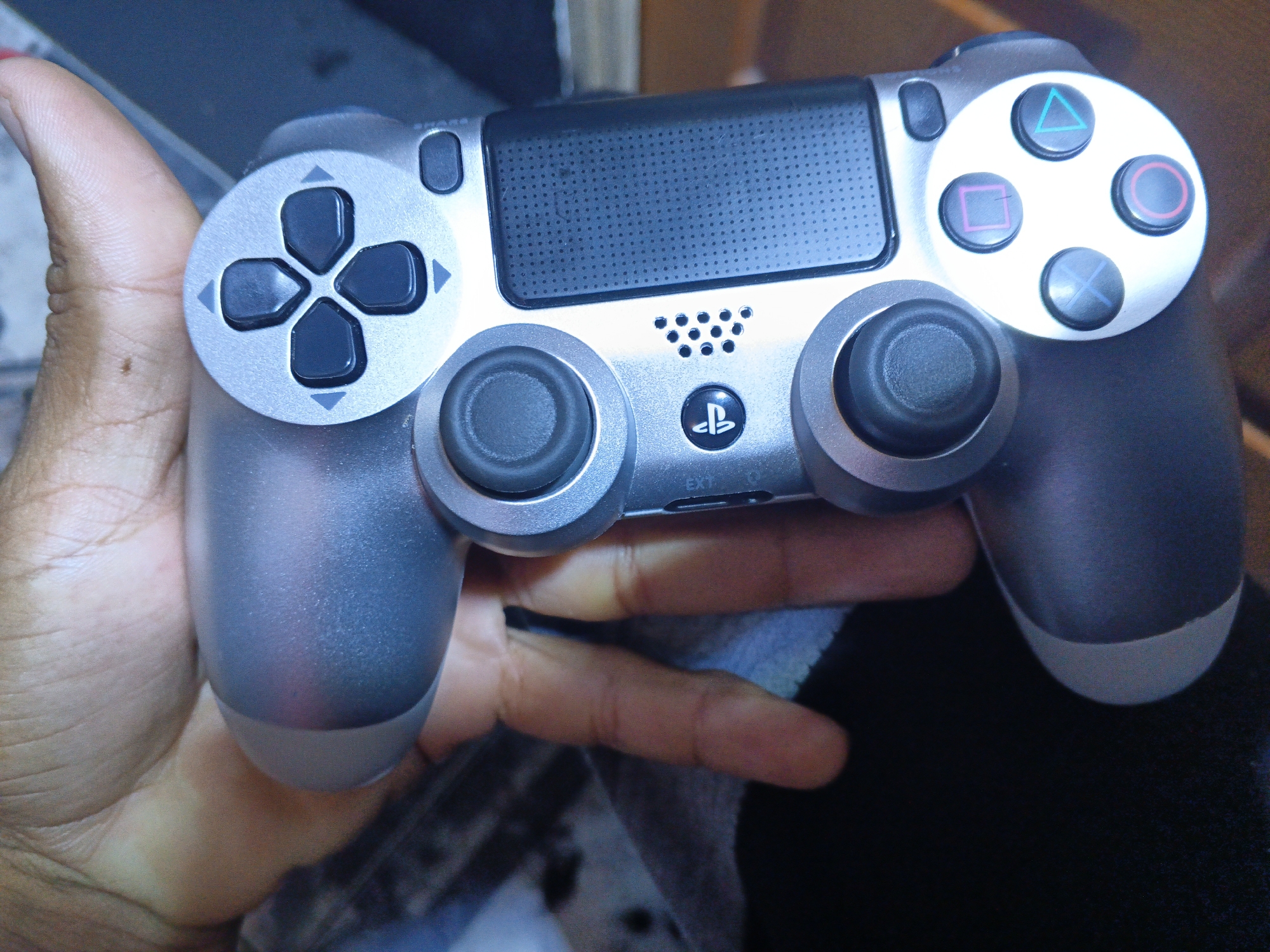 A hand holds a silver Playstation 4 Dualshock controller. The controller is in good condition, with all buttons and analog sticks visible. The location is unknown.