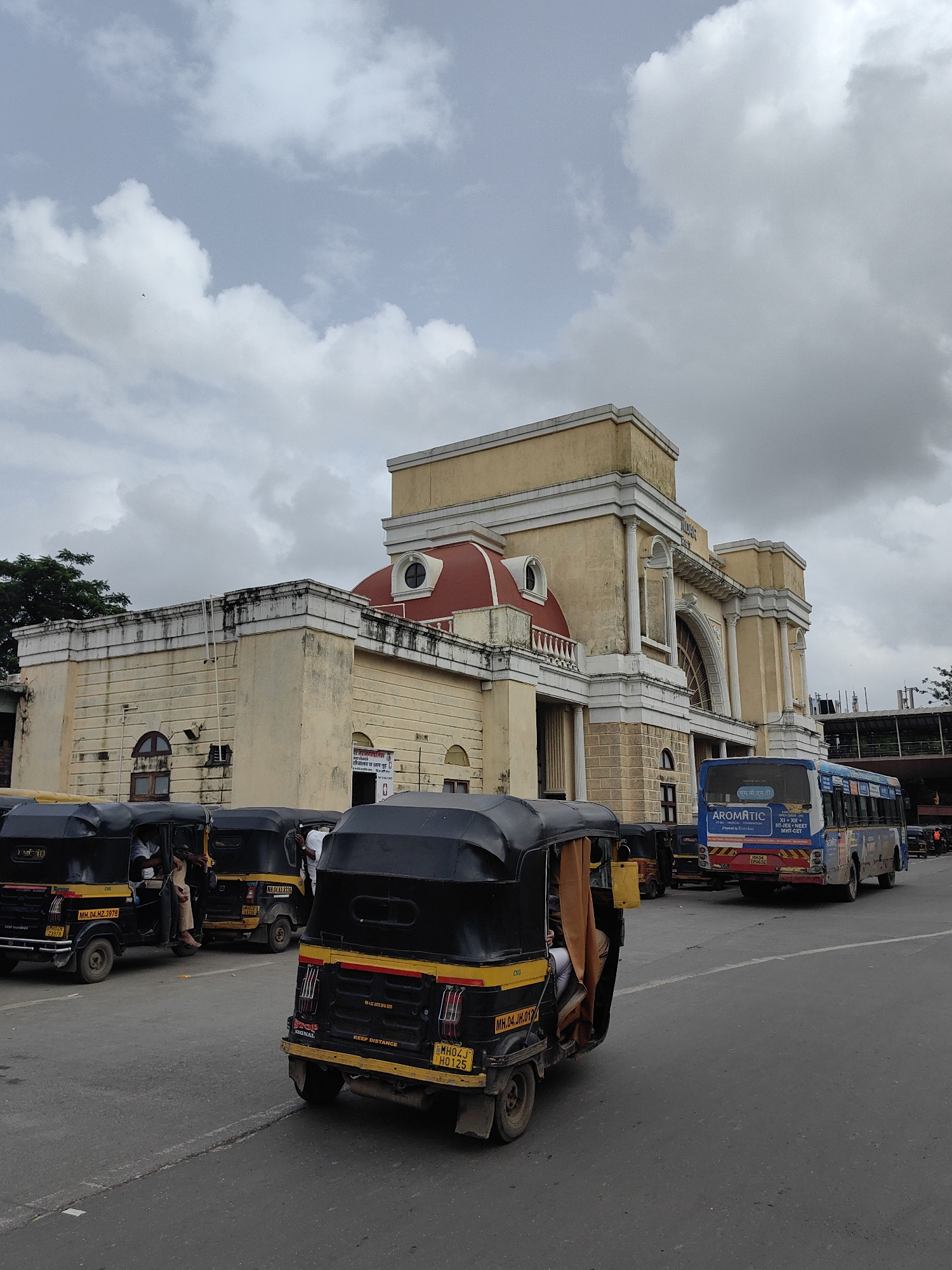 The image depicts a street scene in front of a grand, yellow building with classical architectural elements, possibly a train station or a government building, in Mira-Bhayander, India. The sky is overcast with dramatic clouds. Several black and yellow auto-rickshaws are parked and moving on the asphalt road. One auto-rickshaw in the foreground is facing away from the camera. A blue and white bus with advertisements on its rear is visible behind the auto-rickshaws. People are visible inside some of the auto-rickshaws and the bus. The building features a red dome and ornate columns. The overall atmosphere suggests a busy urban environment on a cloudy day.