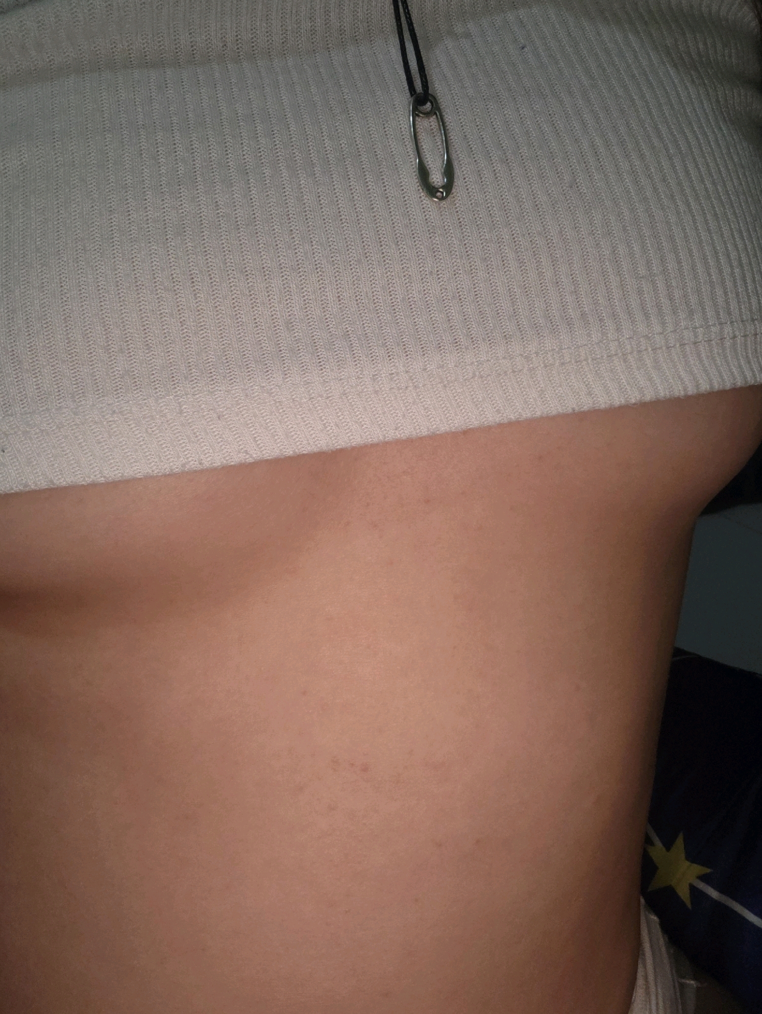 The image displays a close-up, side-view of a person's torso, focusing on the area below the chest and above the waist. The individual is wearing a white, ribbed crop top. A simple necklace with a silver safety pin pendant is visible on the top. The skin tone is fair with some minor blemishes. The background is blurred and appears to be indoors, with a hint of a blue fabric with a yellow star pattern visible on the right side. The lighting is dim, suggesting an indoor setting, possibly a bedroom. There is no discernible information that would place this image in Phan Thiet, Vietnam.