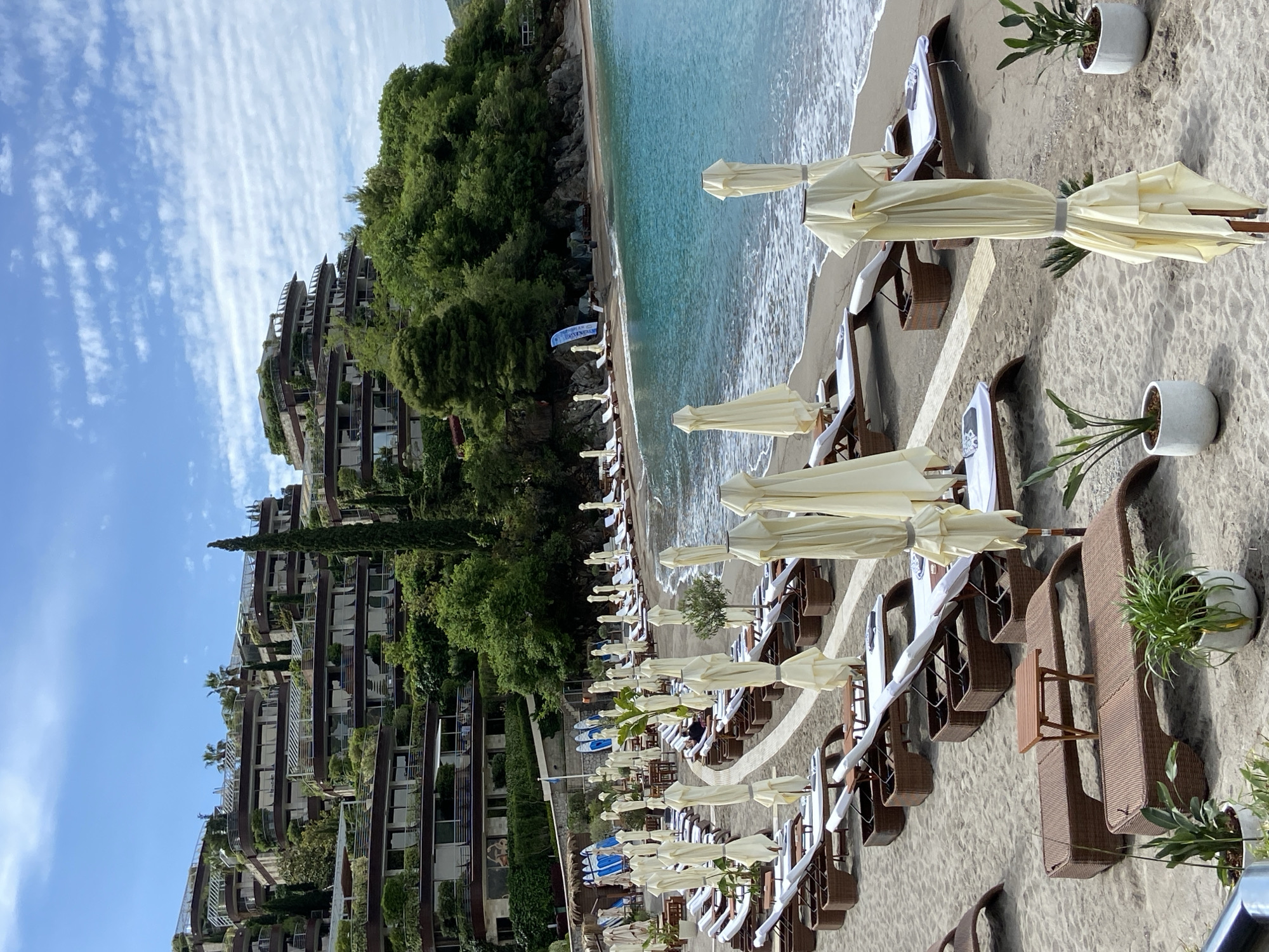 The image depicts a beach in Budva, Montenegro. Rows of lounge chairs with folded white umbrellas are arranged on the sandy beach. Behind the beach are buildings covered in green vegetation. The turquoise sea meets the shore, and a clear blue sky with wispy clouds is visible above. Some of the lounge chairs have small tables and potted plants next to them.