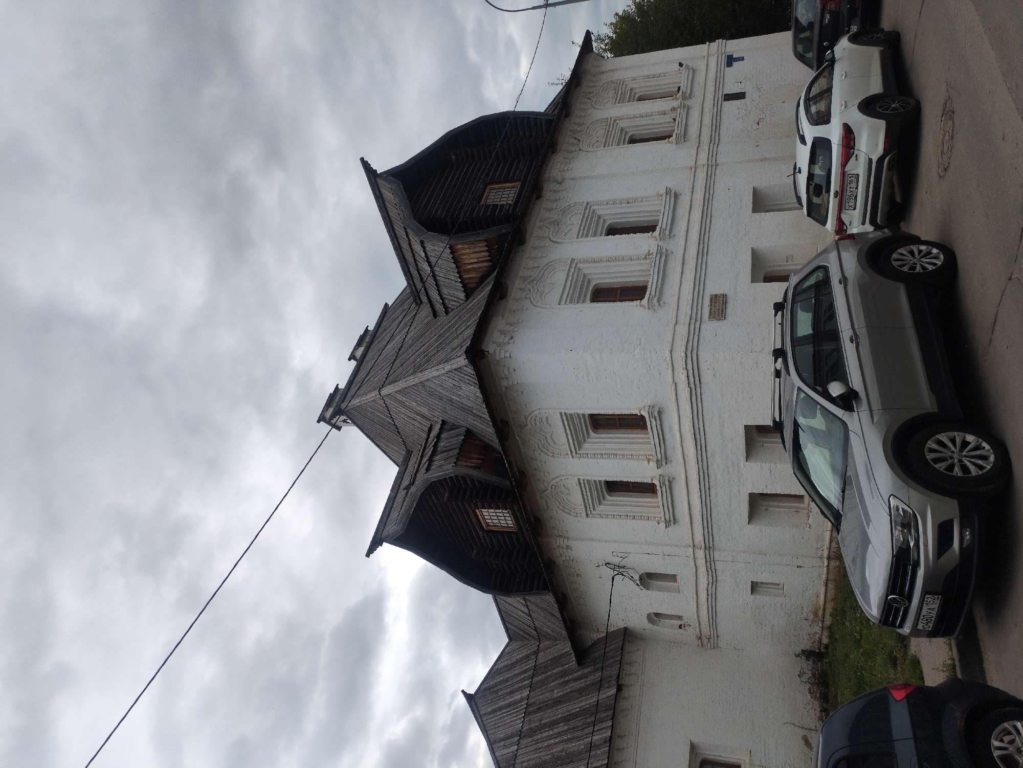 The image shows a white brick building with a complex, multi-gabled wooden roof. The building has several windows, some with decorative white frames. In front of the building, parked cars are visible: a silver Volkswagen Tiguan in the foreground, a white Kia Rio, and a dark-colored sedan partially visible to the right. The sky is overcast and cloudy. A wire runs diagonally across the upper portion of the image. The building is identified as part of the Nizhny Novgorod Kremlin in Nizhny Novgorod, Russia.
