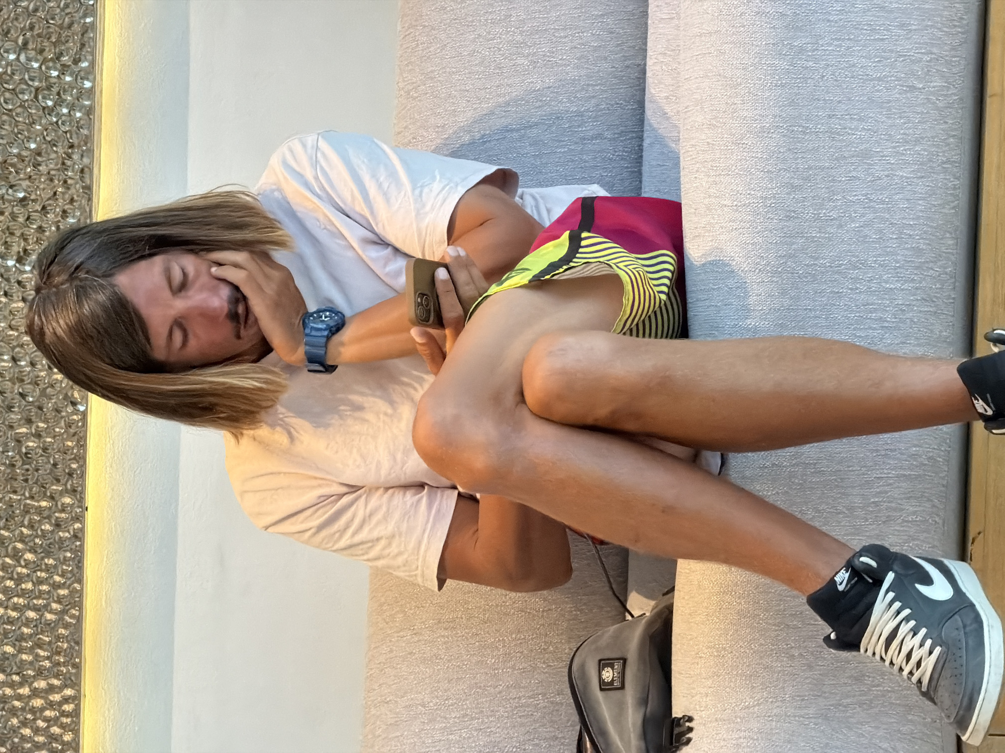 A man is sitting on a light gray couch, looking at his phone. He has long, brown hair and is wearing a light pink t-shirt, multi-colored shorts, and dark gray Nike sneakers. A gray bag is on the couch to his left. The man is resting his head on his hand, and his legs are crossed. The background appears to be an indoor setting with a textured wall and a band of light. This scene is taking place in Sharm El Sheikh, Egypt.