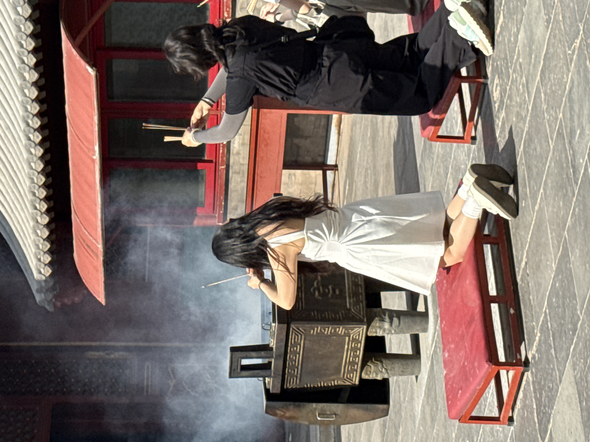 In the courtyard of a traditional Chinese temple, possibly located in Dongcheng District, China, two women are engaged in a religious ritual. The scene is characterized by the presence of incense smoke, a large bronze censer, and architectural elements of the temple.
On the left, a woman with long, dark hair is kneeling on a red cushion. She is wearing a white sundress and white socks with sneakers. Her back is to the viewer, and she is holding an incense stick in her right hand, directing it towards the censer. The censer is ornate, with intricate carvings and lion-shaped feet, and appears to be the source of the rising smoke.
To the right, another woman is also kneeling on a red cushion. She has dark, shoulder-length hair and is wearing dark clothing, including a black top and pants, and a crossbody bag. She is holding two incense sticks in her outstretched hands, also seemingly directed towards the censer. Her focus is directed upwards, possibly in prayer or contemplation.
The ground is paved with stone tiles, and the background features the red and gray-tiled roof of a temple building, with ornate eaves. The overall atmosphere suggests a moment of quiet devotion and reverence.