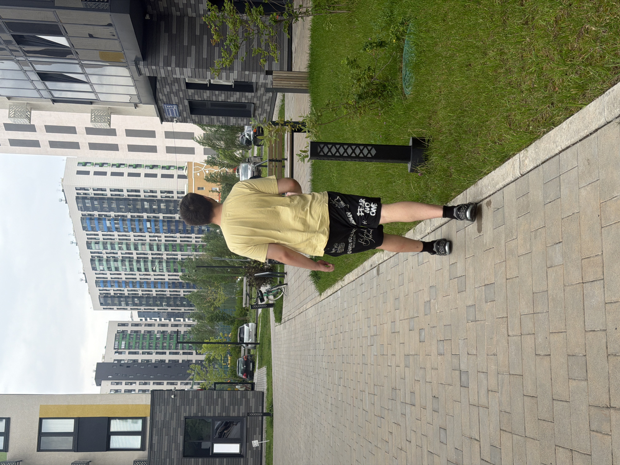 The image shows a young man walking on a paved pathway in an urban environment. He is wearing a yellow t-shirt and black shorts with white lettering. The pathway is made of rectangular beige paving stones and is bordered by a grassy area with a few small bushes and a black light fixture. In the background, there are modern, multi-story apartment buildings with contemporary architectural designs, featuring large windows and balconies. The buildings have a color palette of white, grey, and accents of blue and green. There are cars parked in the distance, and some trees are visible between the buildings and the pathway. The sky is overcast. This scene is consistent with a residential complex in the city of Kazan, Russia, likely a newly developed area with modern infrastructure.