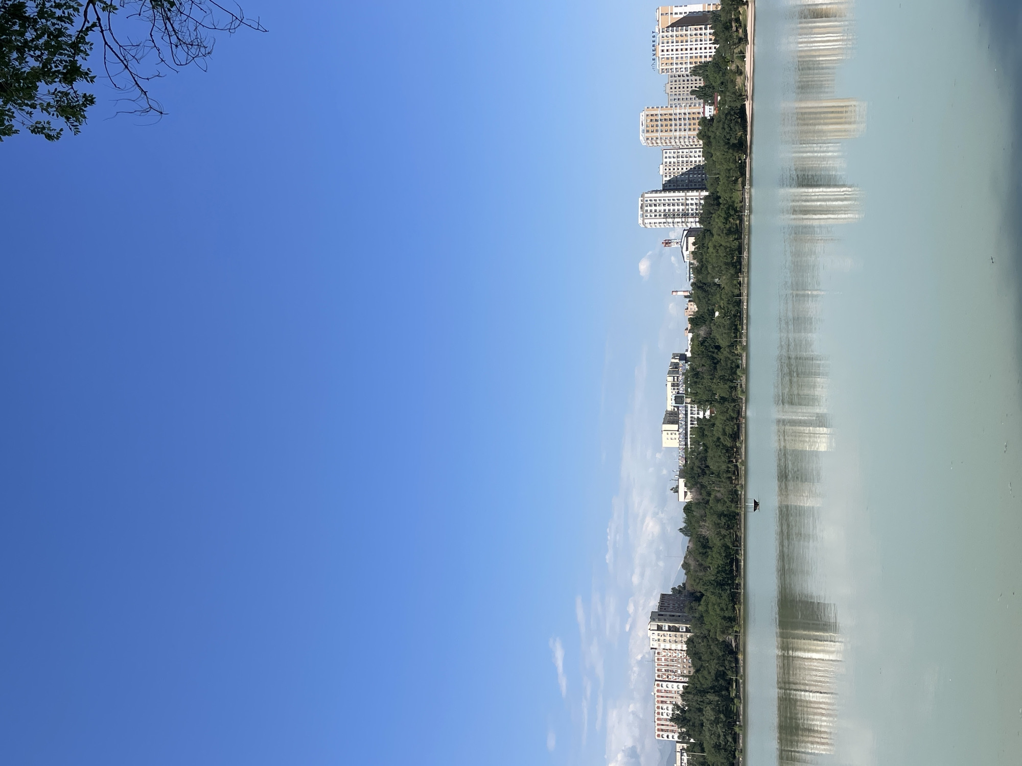 A wide-angle, eye-level shot captures a serene lakefront scene in Almaty, Kazakhstan, likely the Big Almaty Lake. The clear, bright blue sky dominates the upper portion of the frame, with a few wisps of white clouds scattered near the horizon. A lone tree branch with green leaves extends from the upper right corner, framing the expanse of sky.
Below the sky, a line of modern, multi-story buildings stretches across the mid-ground, interspersed with lush green trees. The buildings, primarily residential or office complexes, are a mix of beige, white, and light brown, with visible windows and architectural details. Some of the buildings appear to be taller than others, creating a varied skyline.
The foreground is dominated by a tranquil, light-colored body of water, reflecting the buildings and the sky with remarkable clarity. The water's surface is mostly smooth, with subtle ripples suggesting a gentle breeze. A small, dark, boat-like object is visible in the water, positioned slightly to the left of the center. The reflections create a symmetrical, almost mirror-like effect, doubling the visual presence of the buildings and trees. The overall atmosphere is peaceful and picturesque, indicative of a sunny day in an urban setting adjacent to a natural landscape.