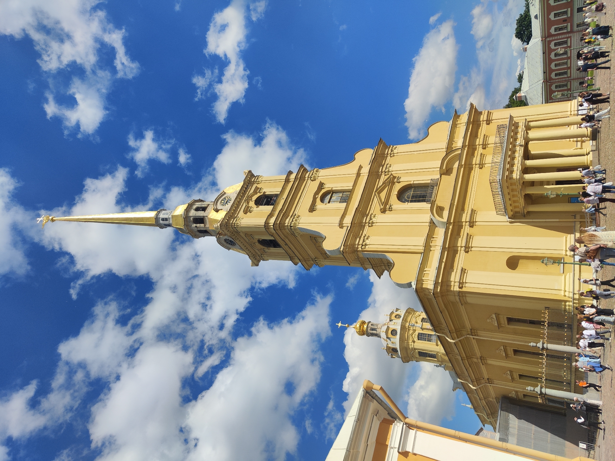 The image captures the Peter and Paul Cathedral, a prominent landmark located within the Peter and Paul Fortress in Saint Petersburg, Russia. The cathedral is characterized by its tall, golden spire topped with a cross, and its bright yellow facade. The sky above is a vibrant blue, dotted with fluffy white clouds, indicating a clear and sunny day.
In the foreground, a paved area is populated by a diverse group of people, suggesting a tourist attraction or public gathering space. The individuals are engaged in various activities, such as walking, standing in groups, and conversing. They are dressed in casual attire, consistent with a warm weather day.
The architectural style of the cathedral is distinctly Baroque, with ornate detailing, columns, and arched windows. The image also shows a portion of another building to the left, also with a light-colored facade, and a red building in the background, which appears to be part of the fortress complex. The overall scene conveys a sense of historical significance and lively public engagement with the landmark.