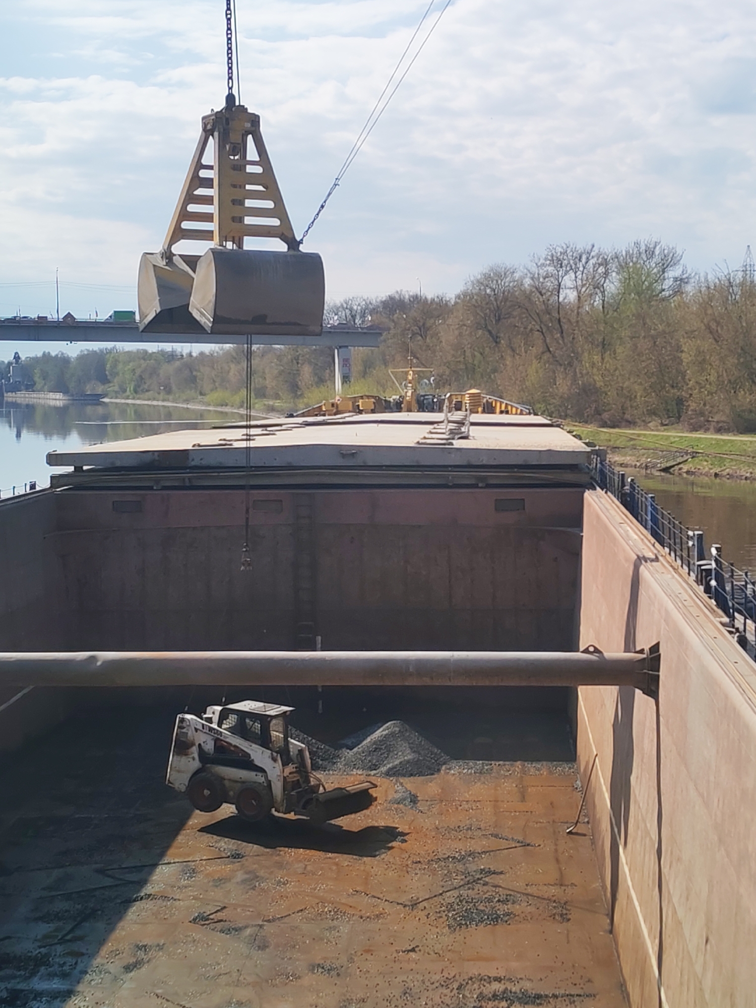 The image depicts a crane unloading gravel or similar material into the hold of a barge on a waterway, likely in the vicinity of Yakhroma, Russia. A skid-steer loader is visible in the hold, possibly used for moving the material once it's unloaded. The barge appears to be positioned near the bank of the waterway, with a bridge visible in the background. The environment suggests a bright, sunny day.