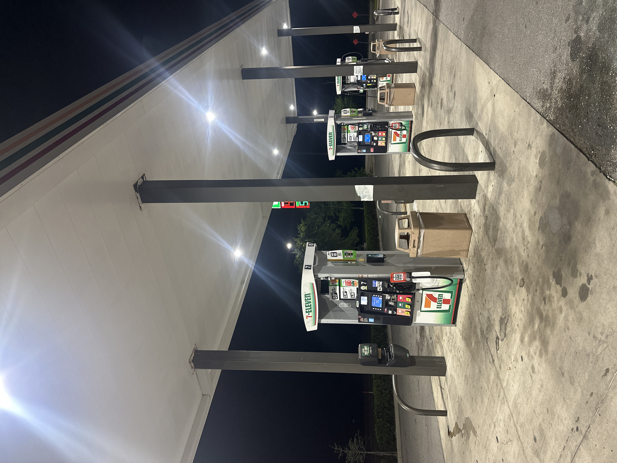 The image depicts a 7-Eleven gas station, likely in Homosassa Springs, Florida, United States. There are multiple gas pumps visible under a canopy, each displaying 7-Eleven branding. The pumps feature digital screens and various buttons. Trash receptacles are positioned near the pumps. Bike racks are present in front of each of the gas pumps. The ground is a concrete pavement. The scene is illuminated by the canopy lighting, and it appears to be nighttime or early morning, as the sky is dark. There are trees and greenery behind the gas pumps.