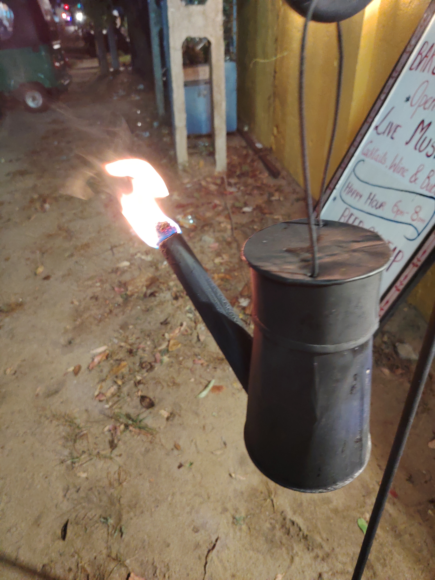 An exterior, eye-level, medium shot captures a black watering can that is emitting a large flame from its spout. The watering can is suspended by a thin metal rod that extends upward and out of frame. The spout of the watering can is long and angled. Behind the watering can, a yellow wall is visible, with a white sign propped against it. The sign has red and blue writing that is partially obscured by the watering can. The ground in front of the watering can is a dirt path with scattered leaves. In the background, a blurred green vehicle with red and white lights is visible on a street. The scene appears to be outdoors at night, possibly in an urban setting, given the presence of the vehicle and streetlights.
