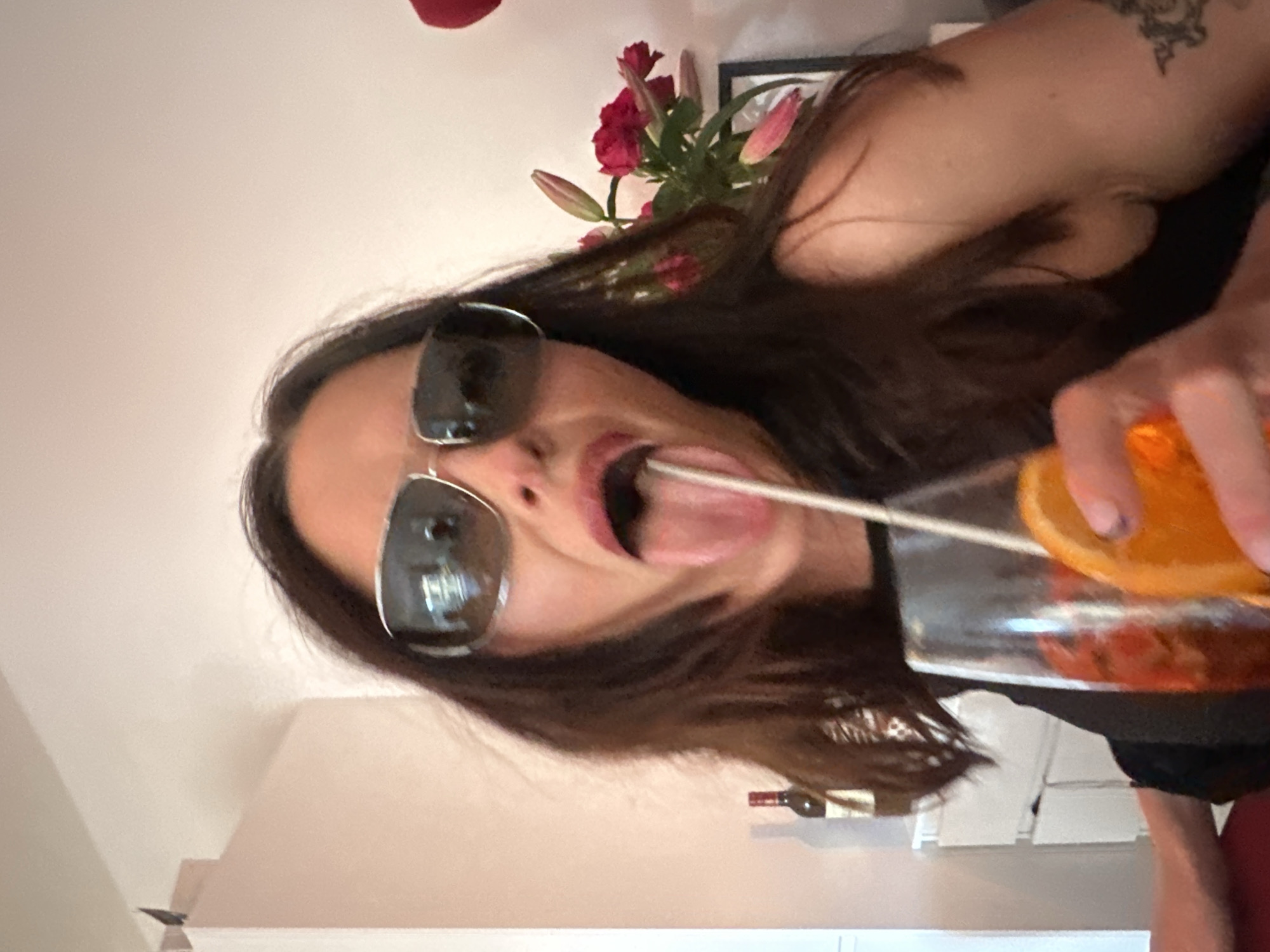 A woman with dark hair, wearing sunglasses, is holding a glass with a straw and an orange slice. She is sticking her tongue out and appears to be interacting with the drink. In the background, there are flowers, a framed picture, a shelf with a wine bottle, and a white wall. The location is not identifiable in the image, and therefore I cannot provide the city or country.