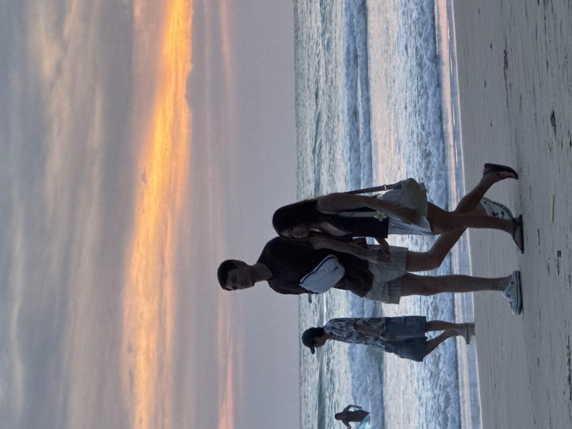 At dusk on Karon Beach in Karon, Thailand, a man and a woman walk hand-in-hand along the shoreline. The man, with dark hair, wears a dark maroon t-shirt and light-colored shorts. A light blue satchel hangs from his left shoulder. The woman beside him has dark hair and wears a dark top and a white skirt. Another man, wearing a patterned short-sleeved shirt and dark shorts, stands to their left, facing the ocean. In the background, to the far left, a person in dark swimwear is partially submerged in the ocean water, holding a surfboard. The waves are breaking on the sandy beach, and the sky is filled with soft orange and grey clouds, indicating the sun has set.