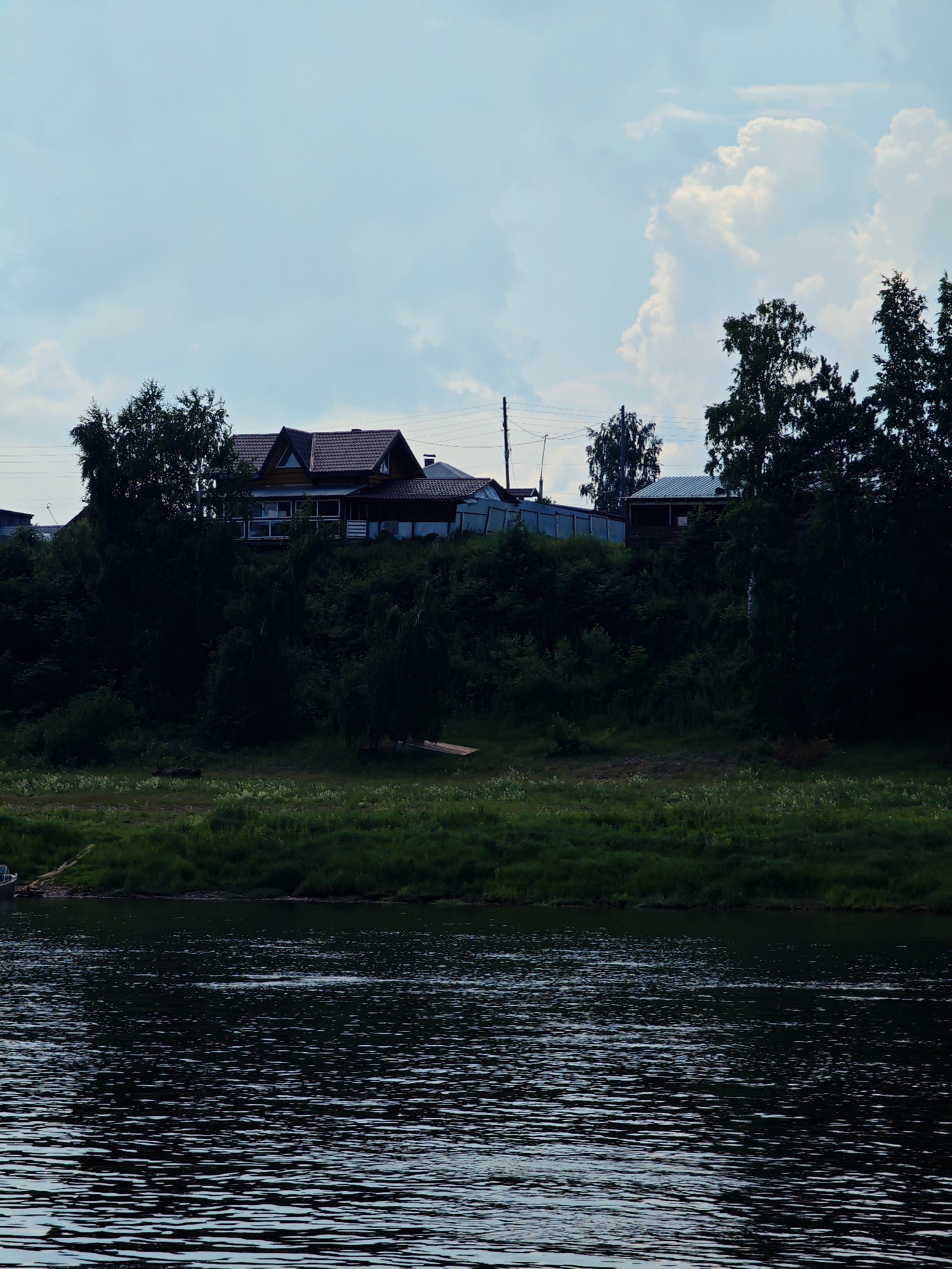 The image shows a wide, dark river in the foreground, with ripples on its surface reflecting the sky. The riverbank on the opposite side is lined with lush green grass. Behind the grass-covered bank, a dense forest of dark green trees covers a steep hill. Atop the hill, silhouetted against a cloudy sky, are several houses with reddish-brown roofs. Power lines are visible strung between utility poles in the background. The overall scene depicts a serene, natural landscape with a few human dwellings integrated into it. The location is identified as Ust-Kem, Russia.