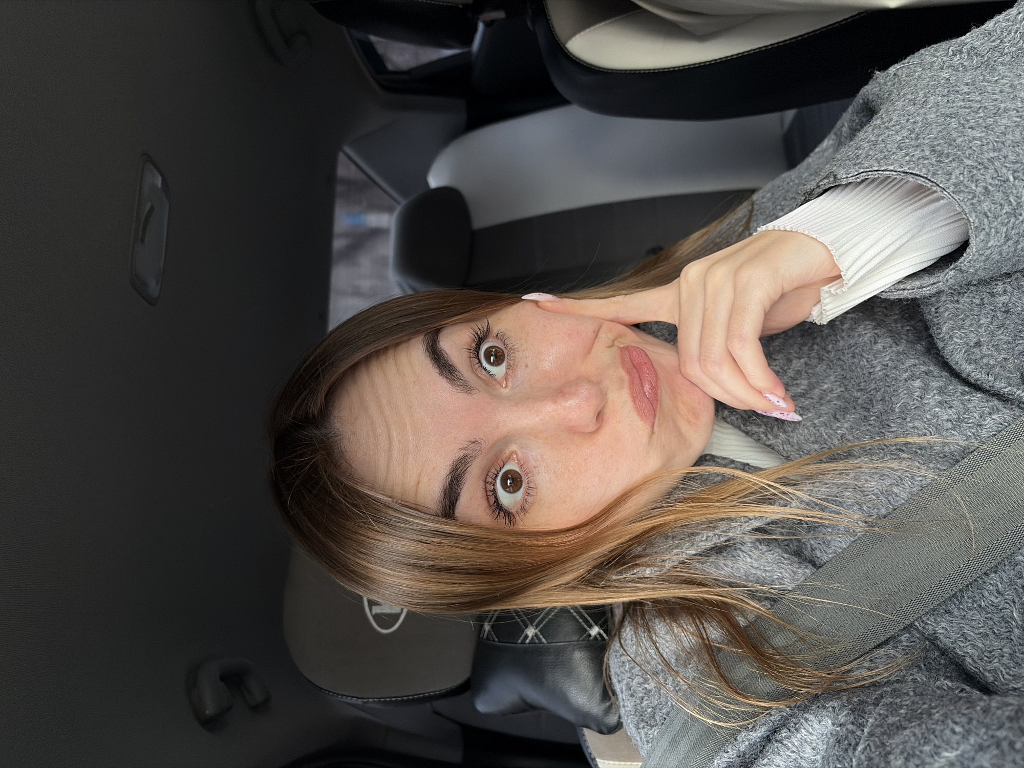 A woman is posing for a selfie in what appears to be the driver's seat of a car. The car's interior is a combination of black, white, and gray. The woman has light brown hair and is wearing a gray coat over a white ribbed shirt. A gray seatbelt is visible across her chest. The lighting suggests she is in an urban setting, possibly Izhevsk, Russia.