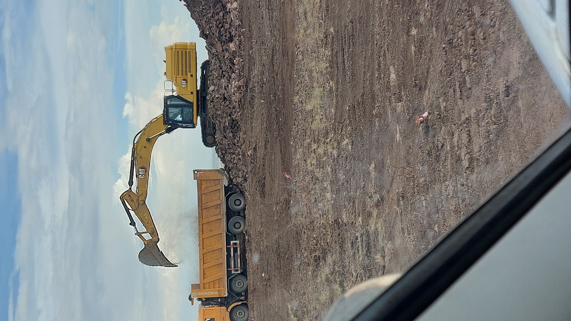 An excavator is dumping rocks into a dump truck on a construction site. The scene is in the city of Saran, Kazakhstan. The sky is cloudy with patches of blue. The ground is dirt and rocks. There is a white vehicle partially in the foreground.