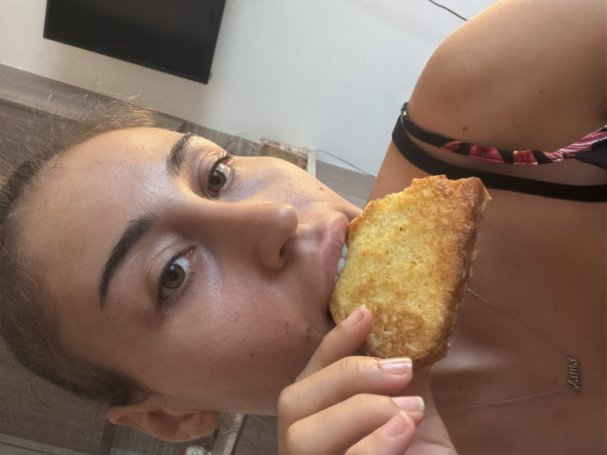 In Dromolaxia - Meneou Municipality, Cyprus, a close-up, medium shot captures a young woman in an indoor setting, with a blurred background suggesting a kitchen or dining area. She is holding a piece of toast to her mouth with her right hand, mid-bite. Her gaze is directed slightly off-camera. She wears a necklace with a pendant that reads "Mama." The environment is casually furnished, with a glimpse of cabinetry and what appears to be a wall-mounted television in the background. The lighting suggests it is daytime.