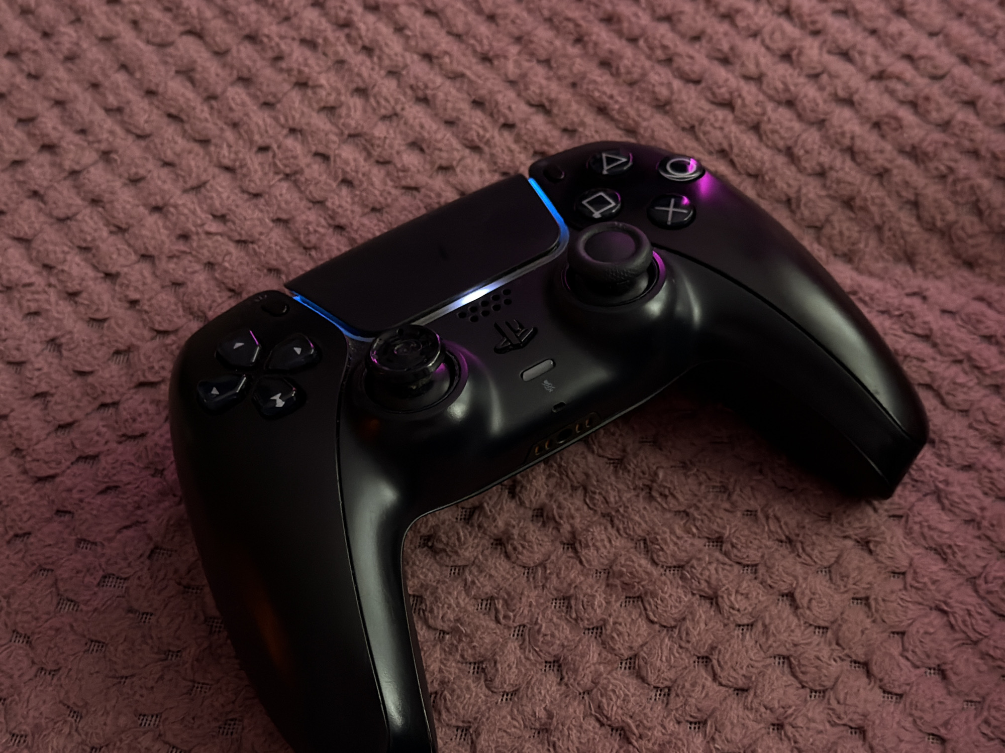 A black PlayStation 5 DualSense controller rests on a textured pink blanket. The controller is positioned diagonally, with the top left corner closer to the viewer. The controller's light bar emits a blue and purple glow. The blanket has a woven pattern of small, raised loops. The scene is an indoor still life.