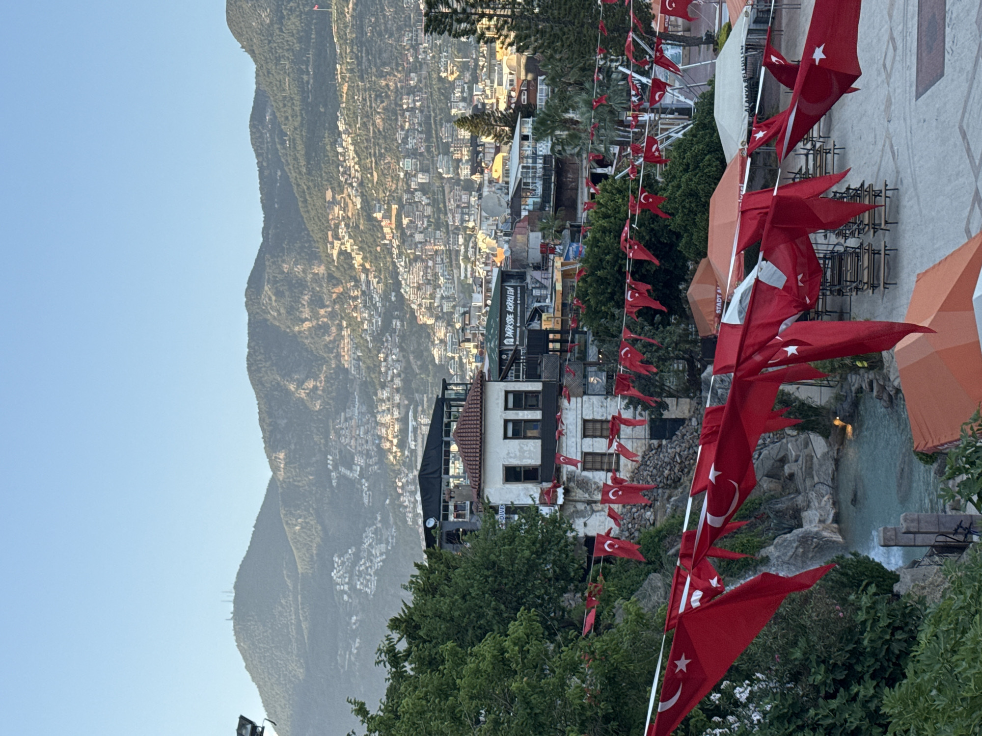 The image shows a view of Alanya, Turkey, from an elevated perspective. A large mountain dominates the background. The foreground includes dense green trees and numerous Turkish flags strung on white poles. These flags are red with a white crescent moon and star. In the midground, a cityscape is visible, with buildings cascading down the hillside. A waterfall or stream flows near the center, and orange umbrellas are visible. The sky is pale blue. The scene conveys a sense of place and suggests a location within a city proud of its national identity.