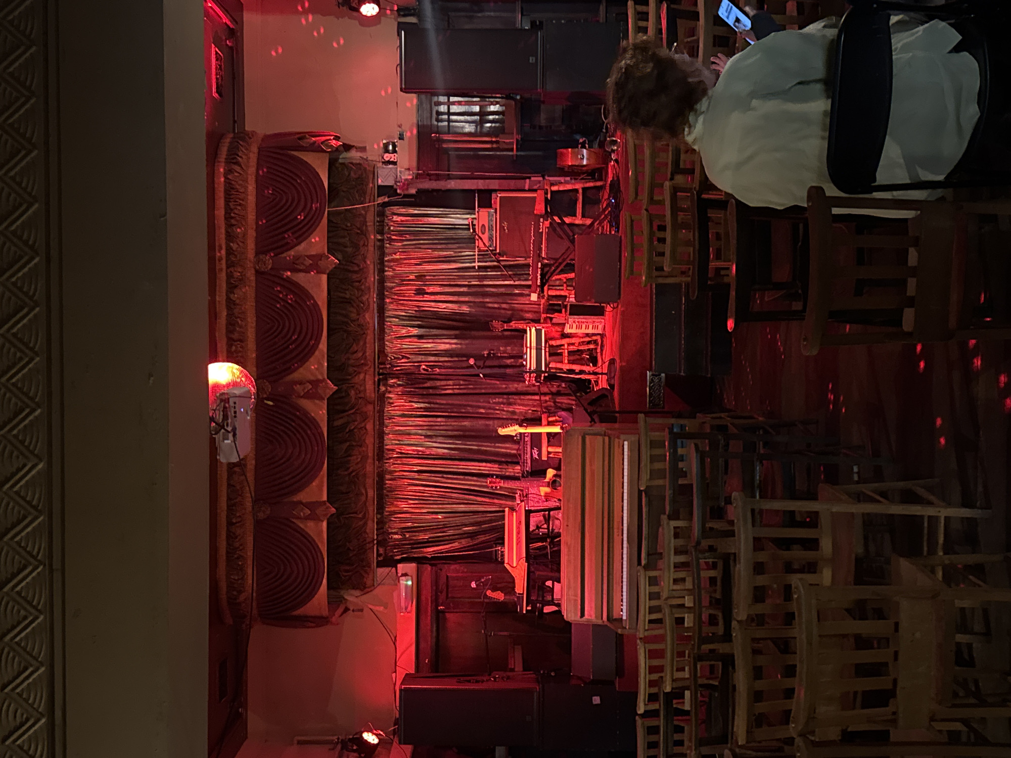 The image captures an indoor scene, likely a music venue or performance space in London, United Kingdom, bathed in dim, atmospheric red lighting. The setting appears to be during the evening or night.
In the foreground and to the right, a person with curly brown hair is seated on a wooden chair, seen from behind. They wear a light-colored shirt and are engrossed in a mobile phone held in their hands. Around them, numerous wooden chairs are stacked or arranged, suggesting either an event is yet to begin, has recently concluded, or is set up for a smaller audience.
The central part of the image features a stage area. A dark, possibly red, curtain serves as a backdrop. Various musical instruments are visible on the stage, including at least two electric guitars, an acoustic guitar, a microphone stand, and a keyboard. The partially visible brand name on the keyboard appears to be "KENZ-T". Several large speaker cabinets and amplifiers are also positioned on or around the stage, indicating a setup for live music. Overhead, spotlights contribute to the red illumination, and small, scattered red dots of light, likely from a disco ball, are visible on the walls and floor.
To the far left, a decorative architectural column with a zigzag pattern adds a classical or ornate touch to the venue. Below this, a long sofa or bench, adorned with multiple rolled, red-patterned cushions, provides additional seating. The overall mood is quiet and anticipatory, typical of a venue before a performance begins or during a lull.