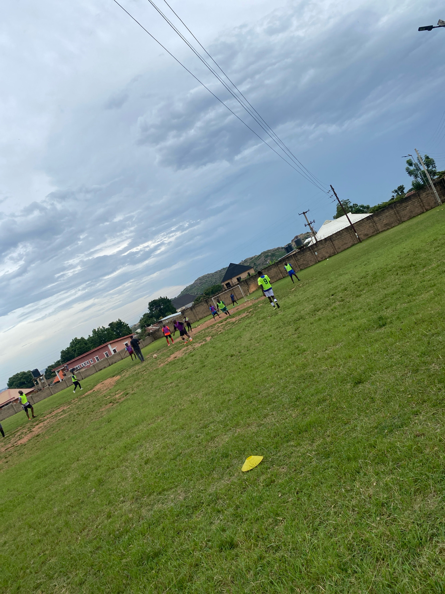 In an outdoor sports field in Jos, Nigeria, several individuals are engaged in a football match under a cloudy sky. The field is covered in green grass, with some patches of dirt visible. A yellow marker lies on the ground in the foreground. In the background, there are buildings and a brick wall, and hills can be seen in the distance. The sky is overcast with a mix of light and dark clouds.