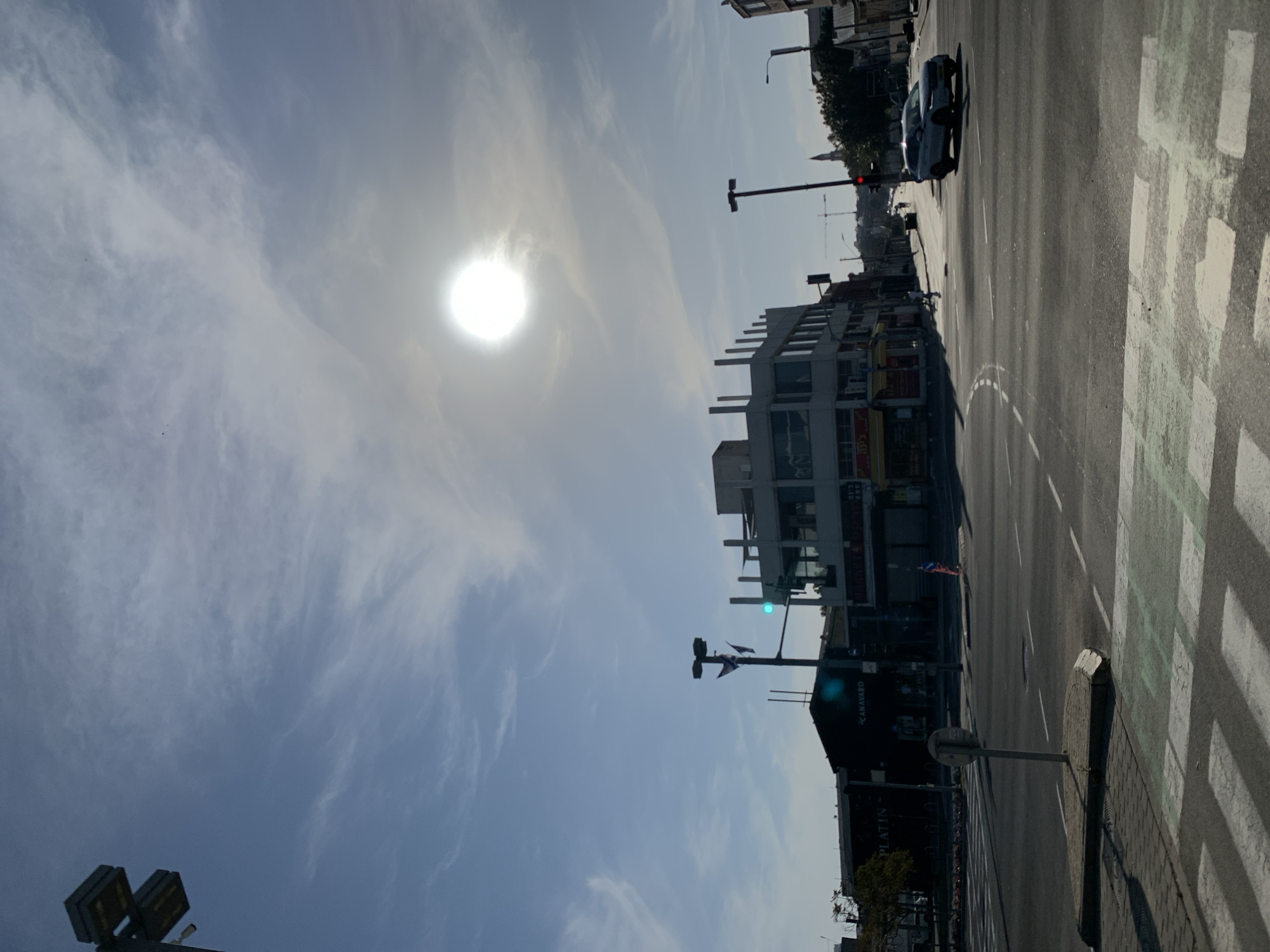 This image depicts a wide-angle view of a street intersection in the Florentin neighborhood of Tel-Aviv, Israel, specifically near Derech Salame 46. The scene is captured under bright, hazy daylight, with the sun prominently visible as a bright disc in the upper-right quadrant of the sky, causing localized overexposure and lens flare. The sky is light blue with scattered, wispy white clouds.
The foreground features an asphalt roadway with white dashed and solid lane markings, alongside a pedestrian crosswalk marked with faded green and white stripes. A small concrete pedestrian island in the mid-foreground contains a vertical metal pole, likely a signpost.
On the left side of the street, a dark-faced building with a "PLATIN" sign is partially visible, followed by a dark commercial building with a segmented roof displaying a white "CANAVARO" sign. At the center of the frame, a multi-story commercial building with a light-colored facade features a distinct upper level characterized by numerous vertical concrete pillars. Its ground floor houses multiple storefronts, some with yellow awnings and visible Hebrew text.
Traffic lights are visible, with a green signal above the central building and a red signal further down the street on the right. A lamppost near the "CANAVARO" building bears two Israeli flags. Further down the street on the right, older, lower-rise buildings recede into the distance, with a distant red spire and a construction crane visible on the horizon.
One individual is observed walking on the sidewalk on the right side of the street, moving away from the camera. A light-colored SUV (possibly silver or light blue) is positioned in the rightmost lane, moving towards the right side of the frame. Minimal vegetation, including some trees and shrubs, is visible along the edges of the frame. Overhead wires are present against the sky.