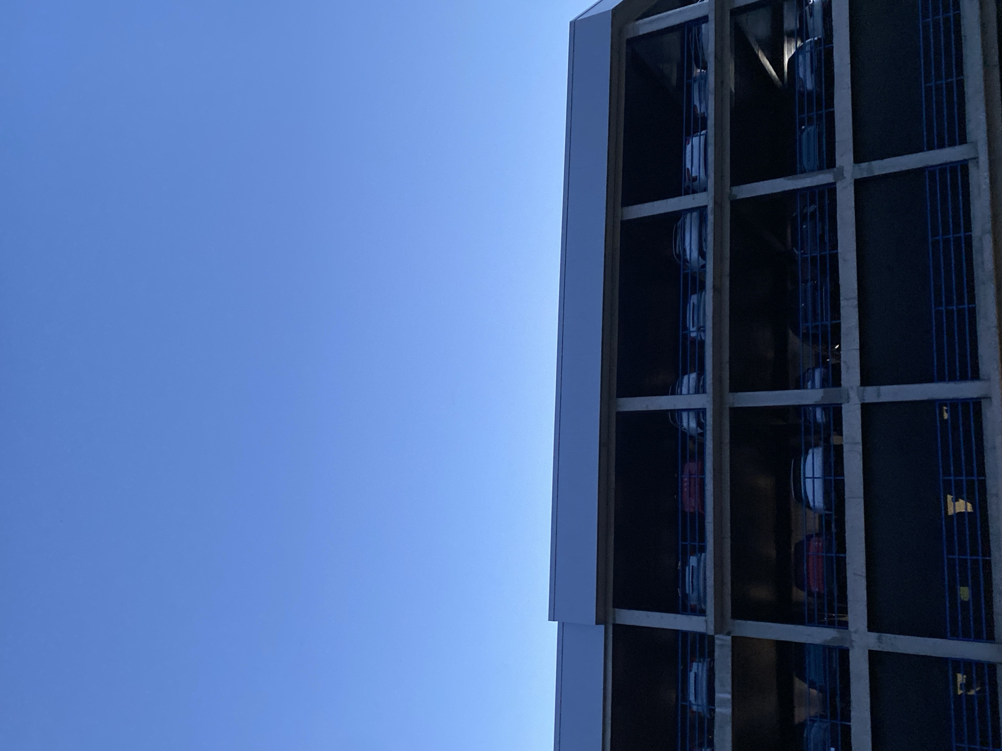 The image shows a section of a multi-level car storage facility, possibly a car park or a dealership's storage lot. The structure is made of concrete and wood beams, with blue metal grates forming compartments for vehicles. Several cars are visible within these compartments, stacked vertically. The cars appear to be white and red, with their tires and some body details discernible. The facility is seen against a clear, bright blue sky, suggesting it's daytime with fair weather. The perspective is from below, looking up at the structure and the sky. The overall scene is urban, with the architectural style of the facility indicating a functional, possibly industrial or commercial setting. There is no visible text in the image.