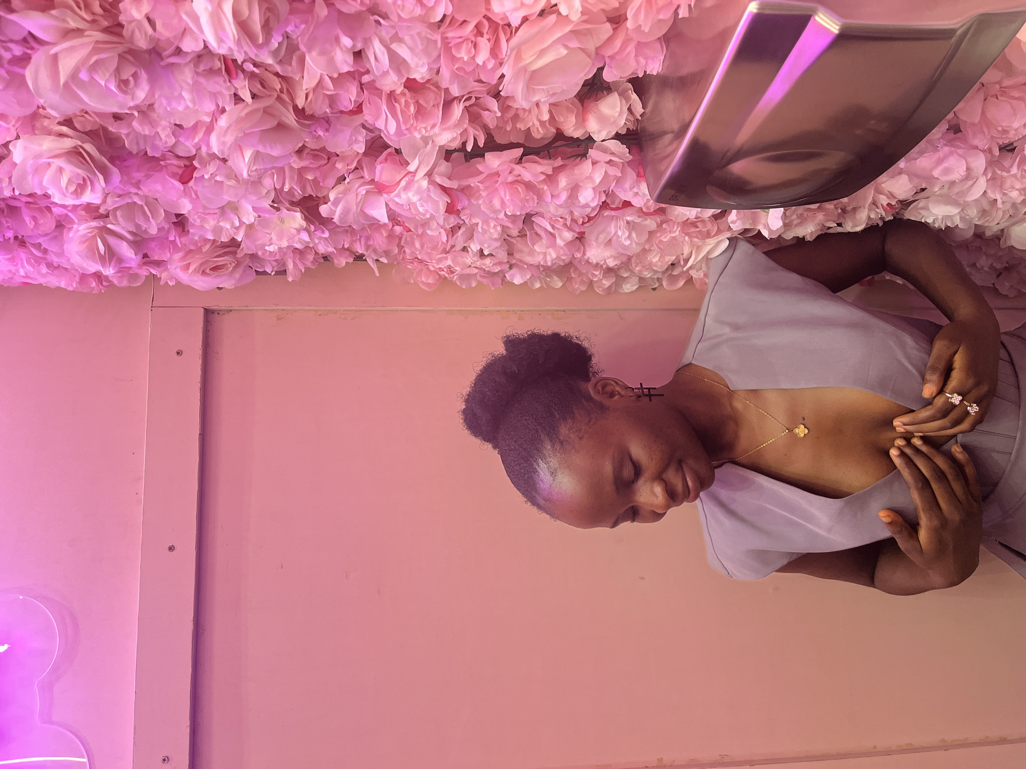 A medium shot captures a young Black woman with her hair in a bun, looking down with a slight smile. She is wearing a low-cut grey sleeveless top, a gold clover pendant necklace, and a ring with two pink flowers on her left hand. She is touching her chest with both hands. To her right, a wall is covered with pink artificial flowers, and a metallic dispenser with pink neon lighting is visible. The background to her left is a plain pink wall, with a hint of a neon light fixture at the top left. The scene appears to be in a decorative, possibly public, indoor space. The location is identified as Howayi, Nigeria.