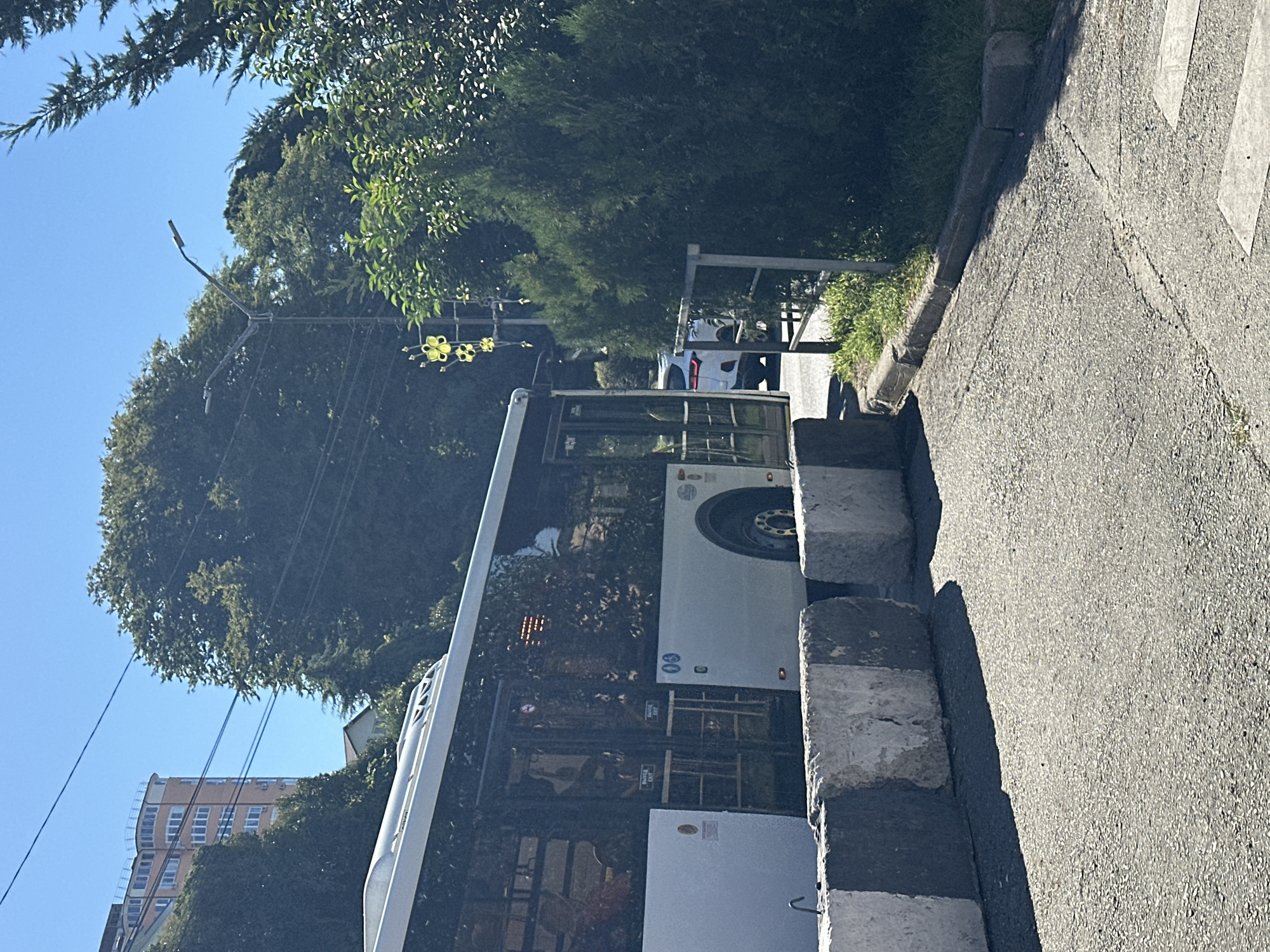 A white public bus is parked on a paved road, partially obscured by concrete barriers. The bus has a modern design with large windows, and some people are visible inside. The scene appears to be in an urban environment, with trees and a building in the background. The sky is clear and blue, indicating a sunny day. The concrete barriers are placed in front of the bus, possibly for traffic control or security. A white car is visible behind the bus on the road. The general location is likely a street or public area in the city of Sochi, Russia, given the presence of public transportation and urban infrastructure. The image captures a still moment with no apparent activity or interaction taking place.