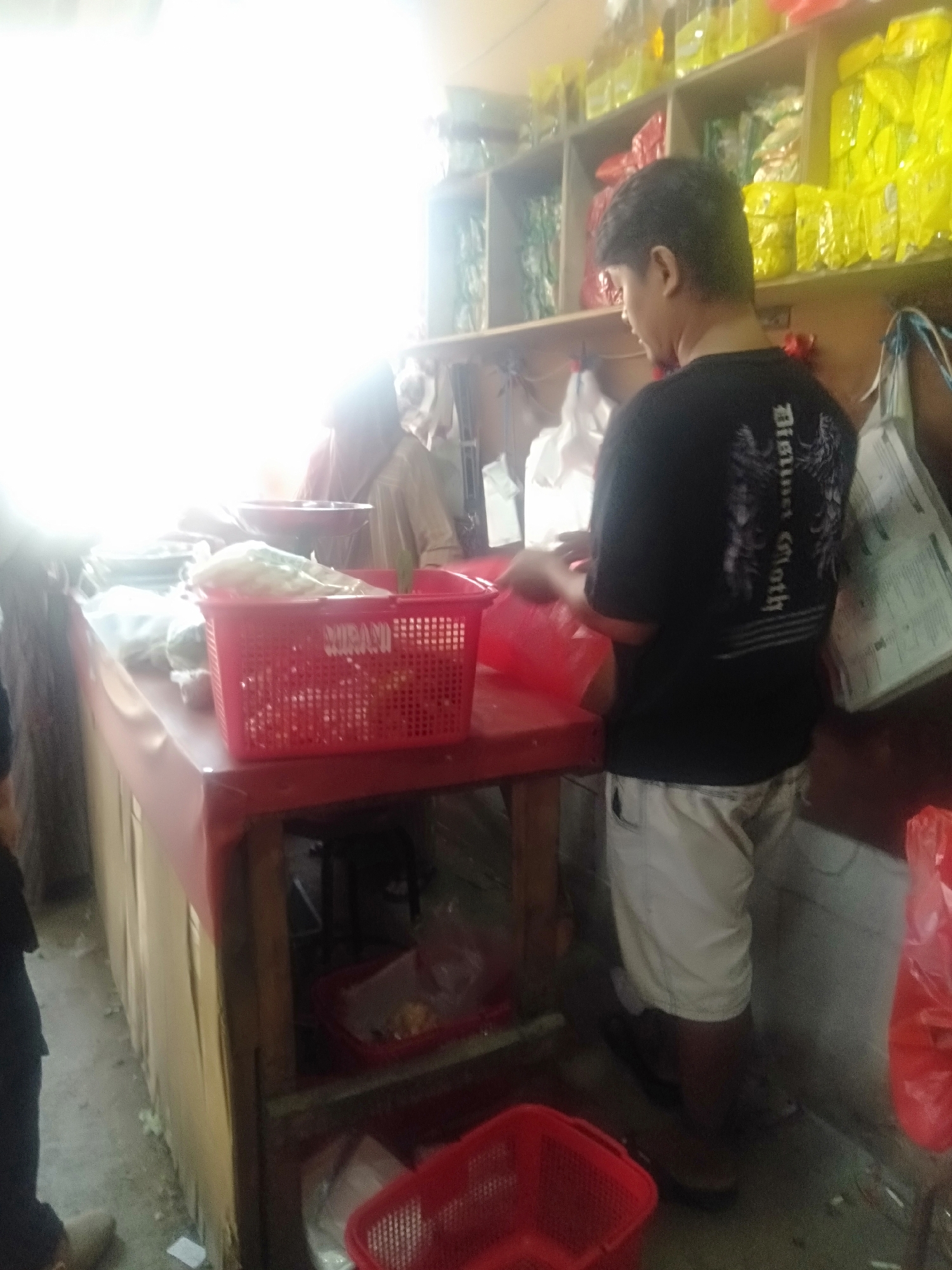 The image depicts an indoor scene, possibly a market stall or small shop, in Sukadami, Indonesia. A young man wearing a black t-shirt with a graphic design and light-colored shorts is standing behind a counter, facing away from the camera. He appears to be engaged in some activity, possibly preparing or packaging items, as his hands are blurred in motion. The counter is covered with a red cloth and displays various items, including red plastic baskets containing food items and plastic bags. Shelves in the background are stocked with yellow and green packaged goods. The lighting is bright and uneven, with a significant glare on the left side of the image, obscuring some details. Another person, partially visible in the background, is wearing a headscarf and a light-colored garment. The overall impression is a busy commercial environment.
