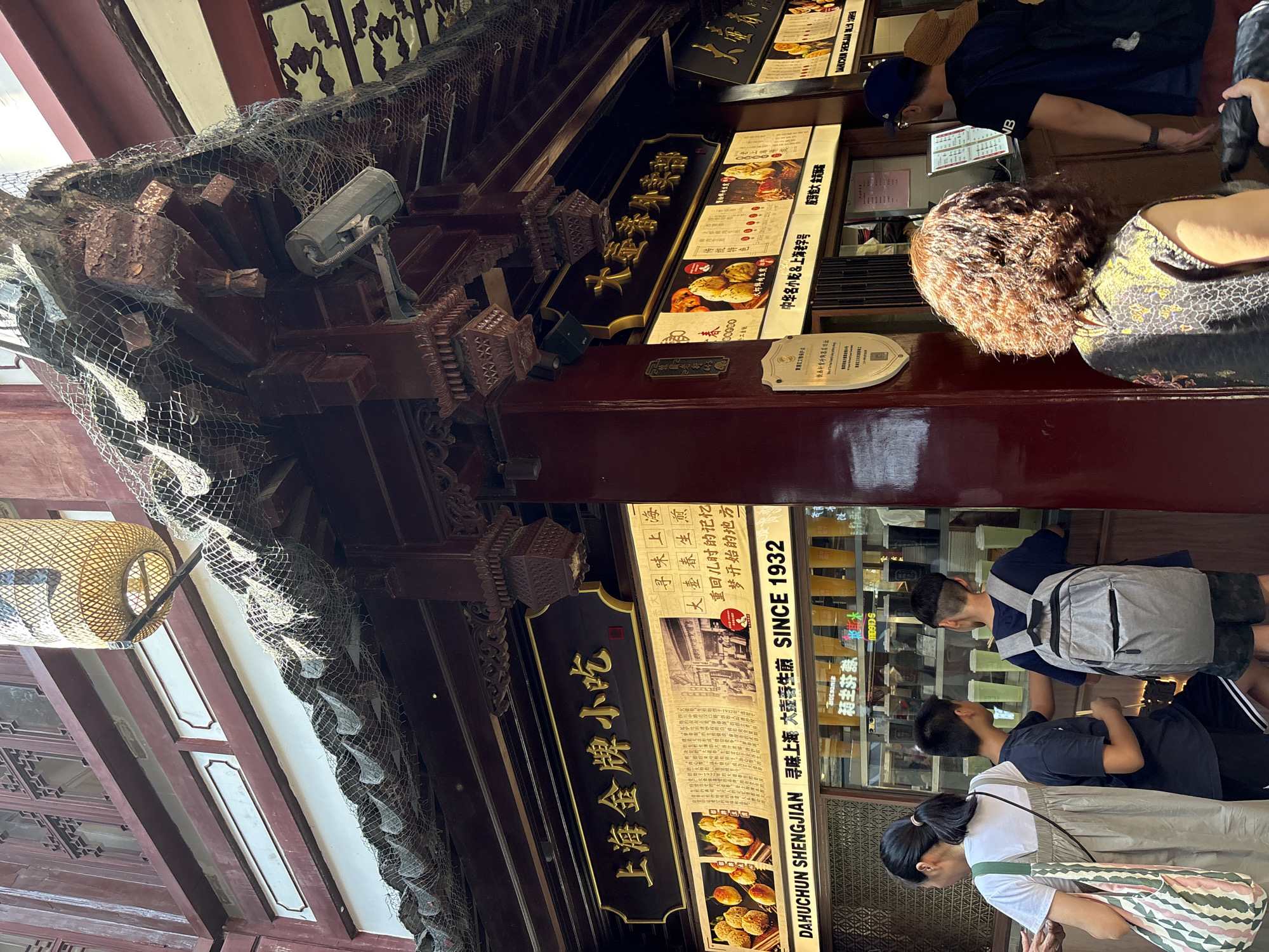 This image depicts the exterior of a food establishment in Shanghai, China, likely a street-facing stall or shop with an outdoor serving area. The architecture is traditional Chinese, characterized by dark red wooden pillars and eaves, intricate carvings, and a tiled roof covered with netting, possibly for protection. A large, woven bamboo lantern hangs from the eaves.
Prominently displayed are signs with Chinese characters in gold lettering against dark backgrounds. One large sign reads "上海金牌小吃" (Shanghai Gold Brand Snacks), and another reads "大壶春鲜肉馅饼" (Dahuchun Fresh Meat Pie). Below these, smaller signs feature images of food, likely the establishment's offerings, and text. One sign prominently displays "SINCE 1932," indicating the business's long history.
Several people are visible, appearing to be customers. In the foreground to the left, a woman with a ponytail wearing a white top and a grey overall dress is holding a striped tote bag. Next to her, two boys are looking towards the serving window; one in a dark t-shirt and shorts is leaning forward, and the other, wearing a backpack, is standing. To the right, a person with curly brown hair in a patterned top is facing away from the camera, standing near a serving counter. Another person in a blue t-shirt and a blue baseball cap is also at the counter, facing the interior of the establishment.
The serving area has a window where beverages in clear plastic cups with yellow lids are lined up. The overall scene suggests a bustling food stall catering to pedestrians in a tourist or commercial area of Shanghai, given the traditional architecture and the type of food being sold.