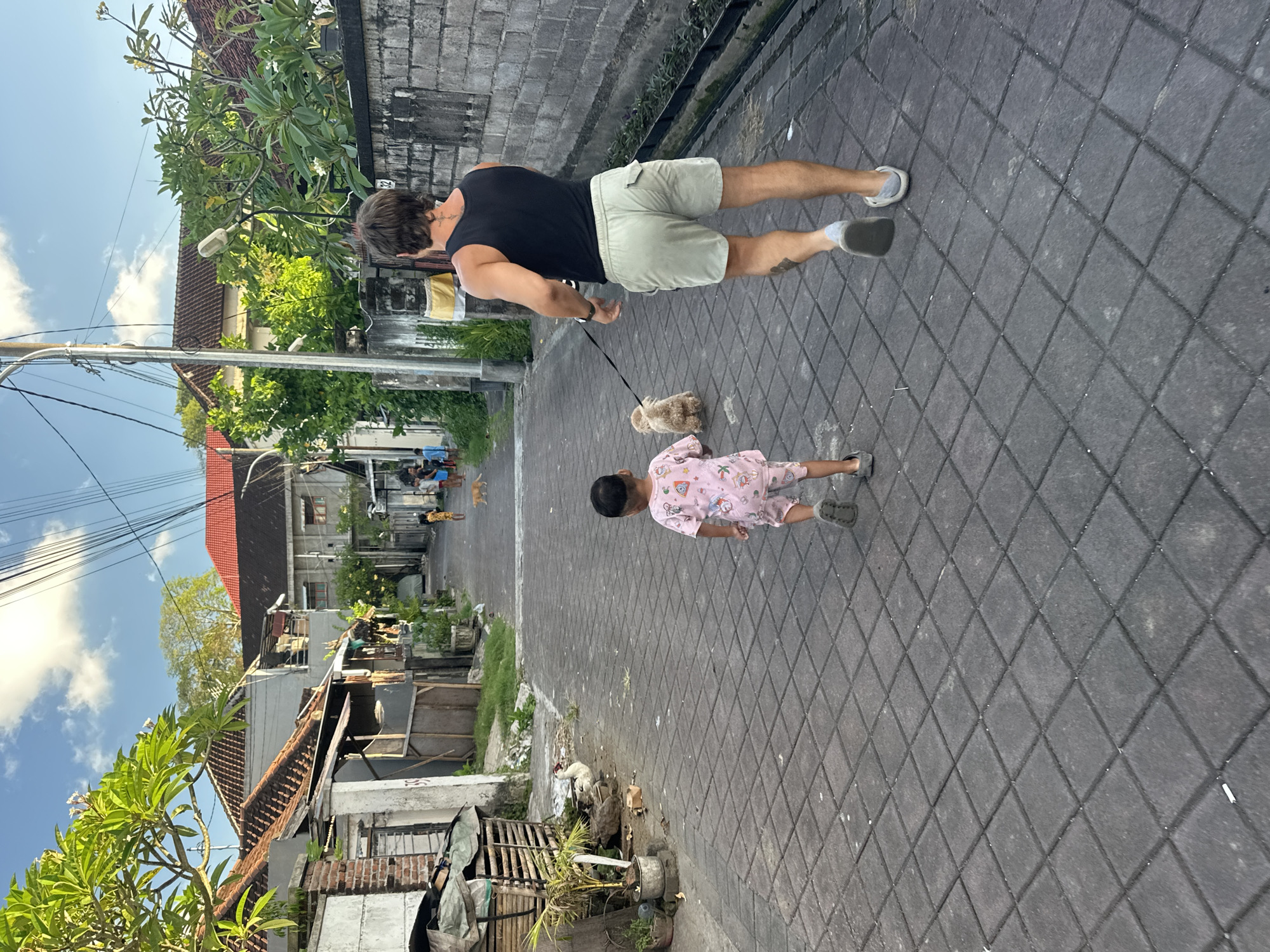 The image captures a scene on a paved street in what appears to be a residential area in North Kuta, Indonesia. A man in a black tank top and khaki shorts is walking a small, tan-colored dog on a leash. A child wearing pink pajamas is walking alongside the dog. The street is lined with buildings of varying styles and conditions. Trees and vegetation are interspersed along the street, adding to the tropical environment. In the distance, several people and dogs are visible on the street. The sky is partially cloudy.