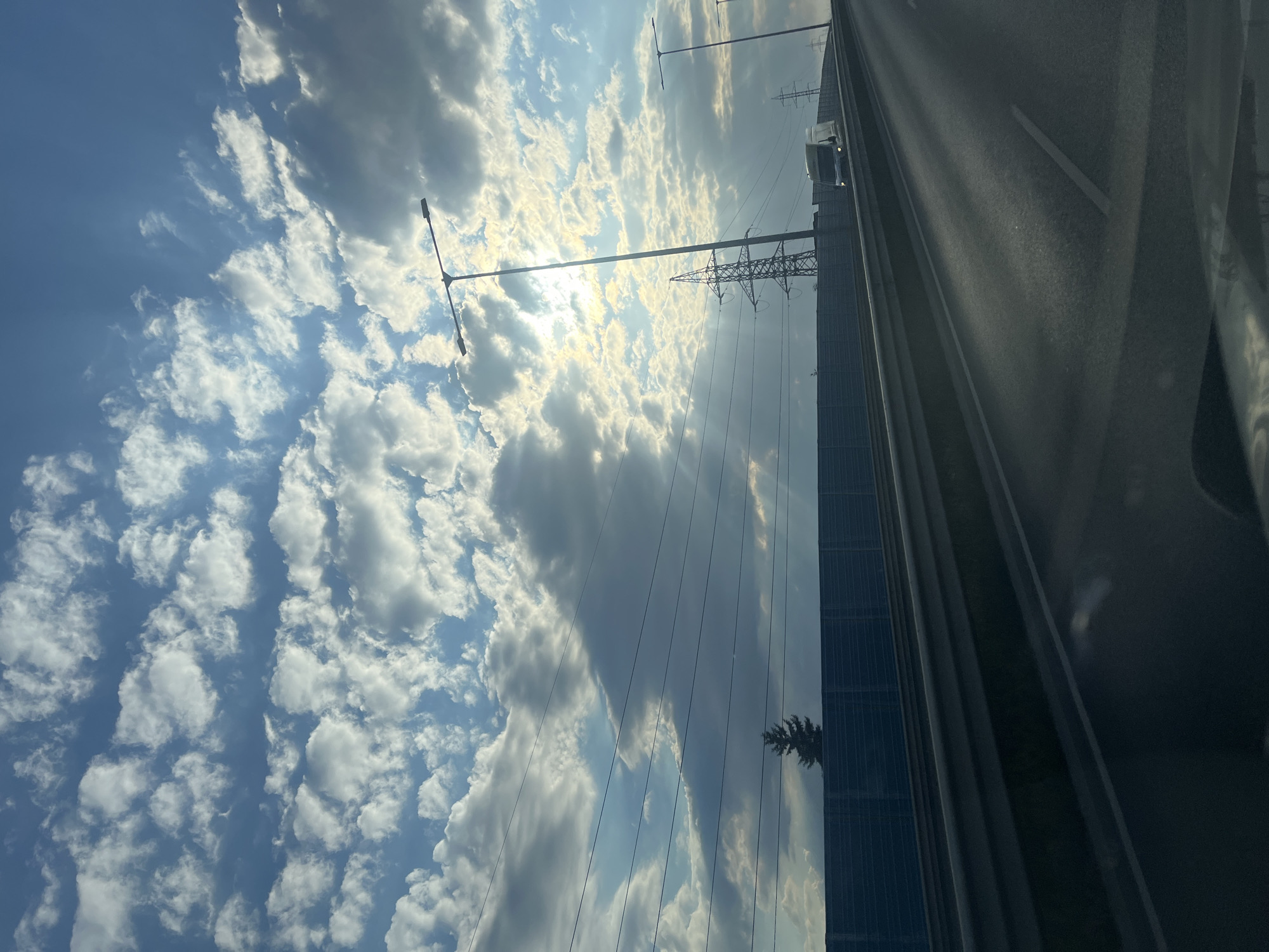 The image shows a view from a vehicle driving on a highway under a cloudy blue sky. The sun is partially obscured by clouds, casting rays of light. On the left side of the highway, there is a tall dark green sound barrier with a single evergreen tree visible above it. Power lines stretch horizontally across the frame above the barrier. Further down the highway on the right, there are streetlights and electricity pylons. A white truck is visible on the road in the distance on the right. The road surface is asphalt with lane markings. The overall impression is a late afternoon or early morning drive, with dramatic cloud formations and the presence of infrastructure.