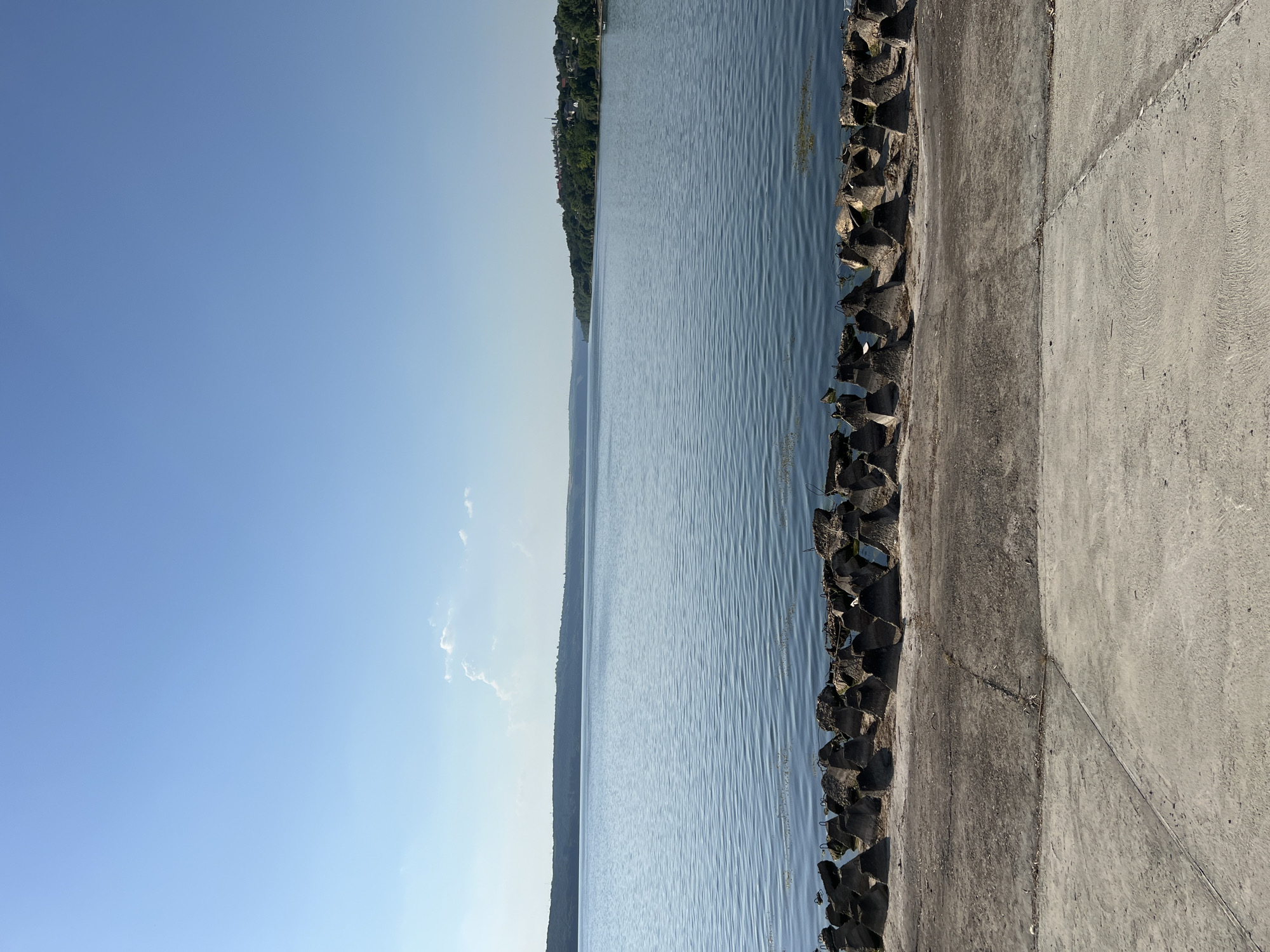 The image depicts a calm, wide body of water, likely a river or a large lake, under a clear blue sky with a few wispy clouds. In the foreground, there is a concrete embankment followed by a line of rough, angular concrete barriers, possibly for erosion control or breakwaters. The water exhibits gentle ripples and reflects the sky. On the opposite shore, a tree-lined hillside is visible, with some buildings and structures forming a small settlement nestled among the trees on the right side of the frame. The overall scene conveys a serene, natural environment with man-made elements at the water's edge. The location is the Nistru River in Korzhevo, Moldova.