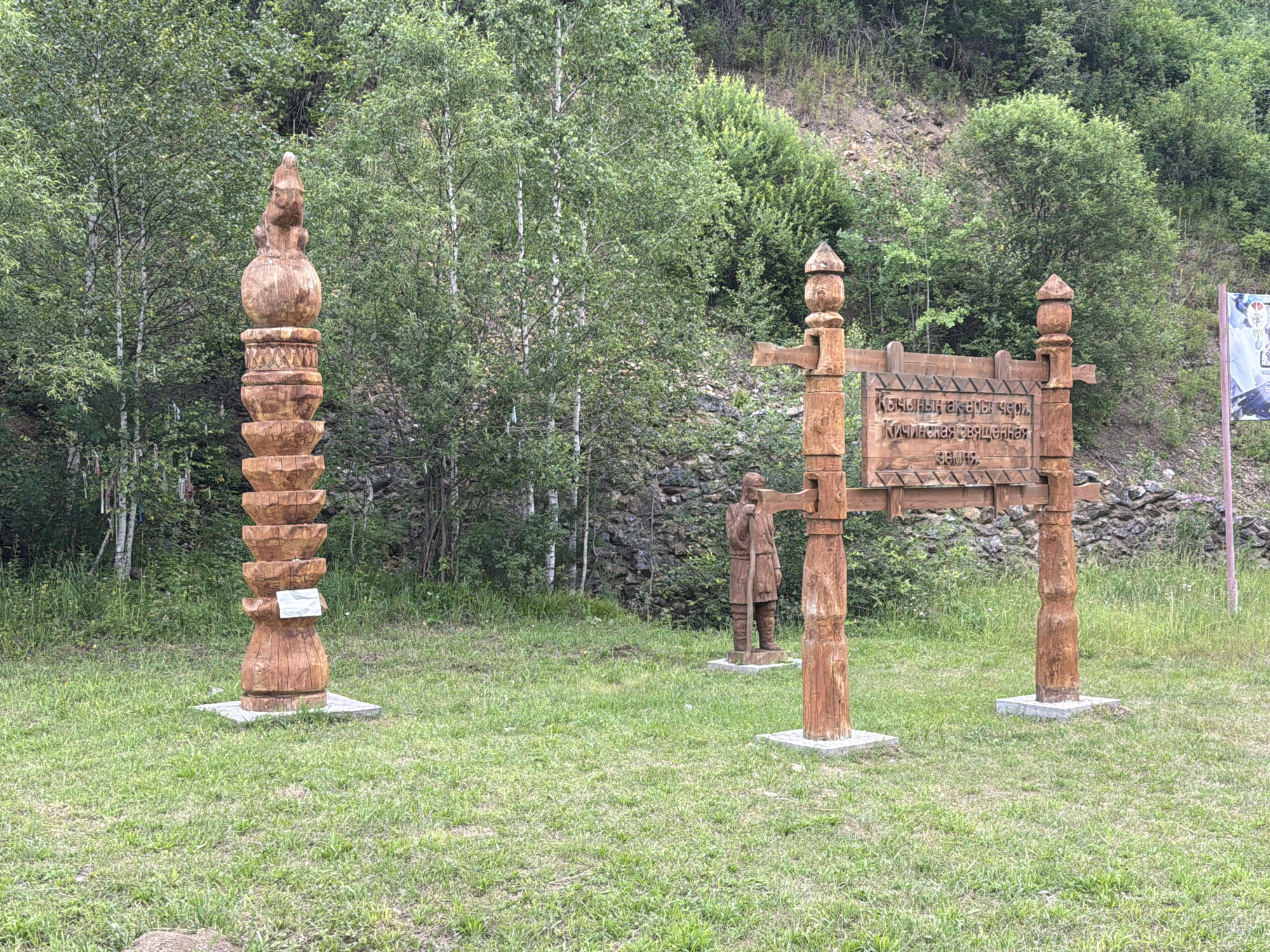 In Srednie Kichi, Russia, an outdoor scene displays cultural carvings. On the left, a tall, intricately carved wooden totem stands on a grey concrete base. To the right, a wooden archway forms an entrance. Two vertical posts, each topped with a carved finial, support a horizontal beam. Mounted on this beam is a wooden sign with text, and to the left of the arch, a wooden statue of a person holding a staff stands on a concrete base. The area is covered in green grass, with a dense forest of trees and a rocky hillside in the background. The sky is overcast.