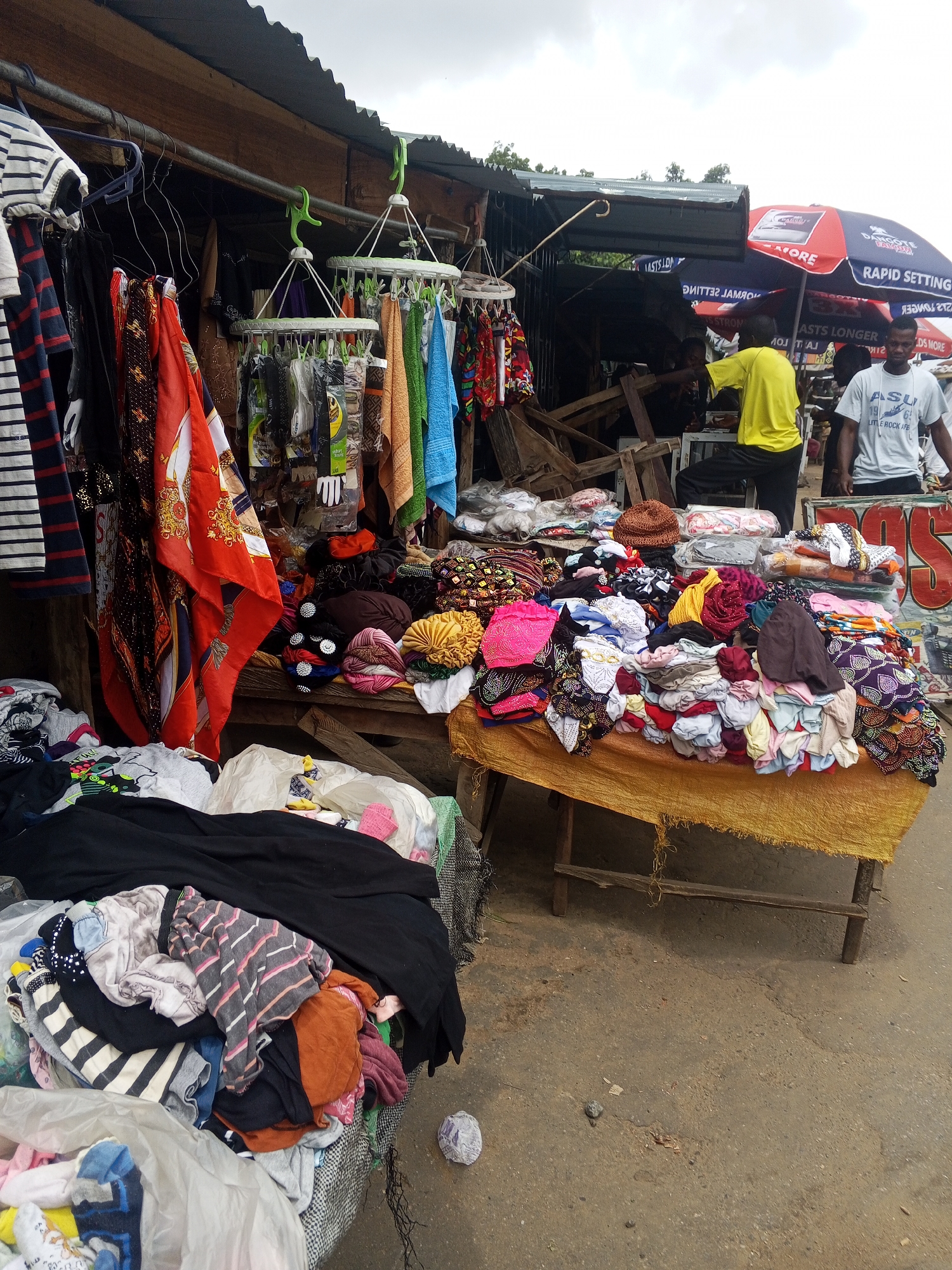 The image depicts an outdoor market scene, likely in Okene, Nigeria. In the foreground, piles of used clothing are displayed on the ground and on a rough wooden table covered with a yellow cloth. The clothing includes t-shirts, dresses, and what appear to be scarves or wraps with vibrant patterns. Behind the table, a stall structure with a corrugated metal roof is visible. Various items are hanging from a rod above the stall, including more clothing and some packaged goods. Two men are present in the background. One man, wearing a yellow t-shirt and black pants, appears to be attending to the stall. Another man, wearing a white t-shirt, stands near a large umbrella that has "RAPID SETTING" and other text on it. The ground is paved with asphalt. The overall impression is that of a bustling, informal market.