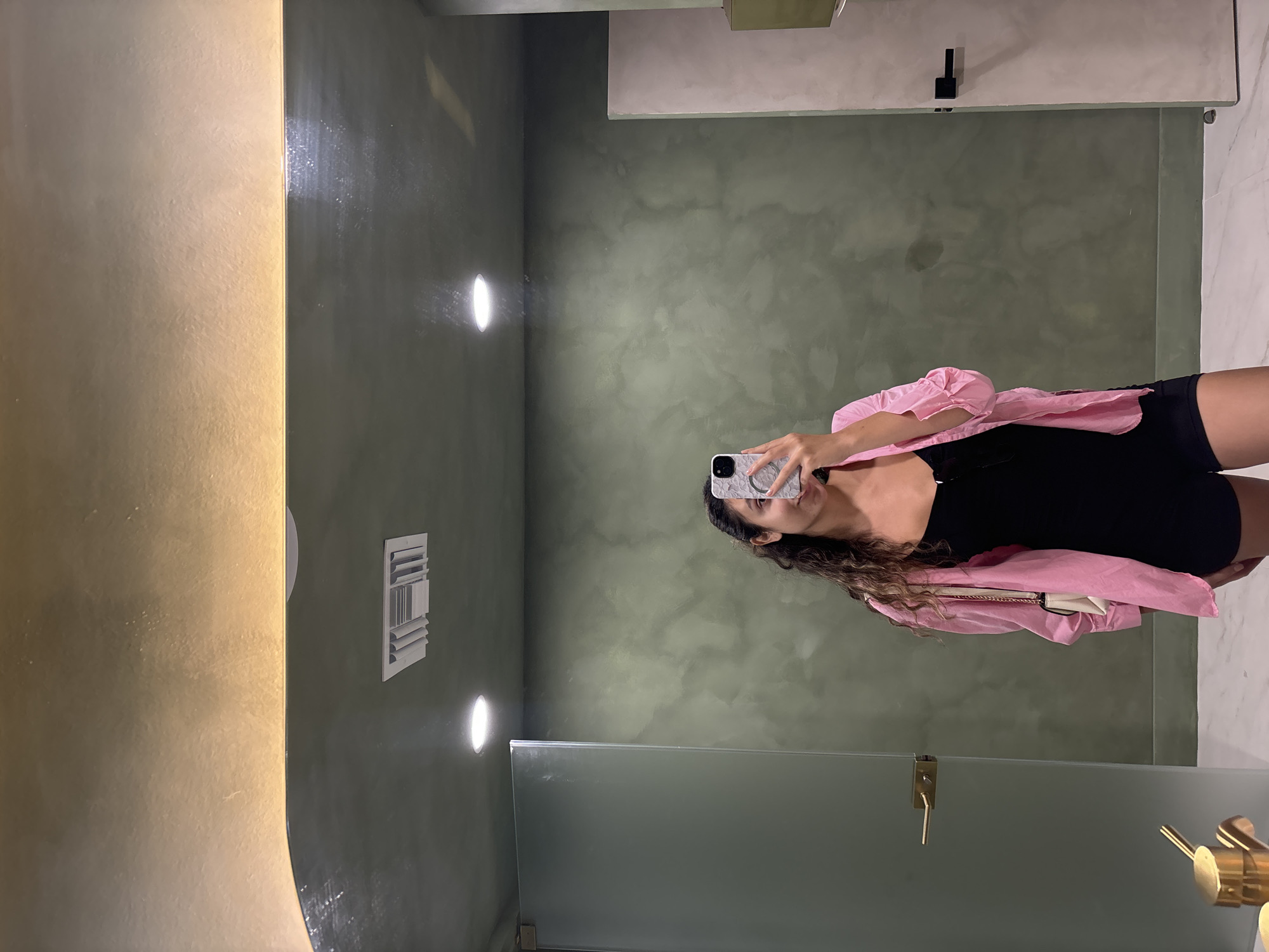 A young woman with dark curly hair stands in a restroom, taking a mirror selfie. She is wearing a black sleeveless bodysuit and a pink open button-up shirt. A small white purse with a gold chain strap is visible over her left shoulder. She holds a smartphone in her right hand, using its camera to capture her reflection. The restroom features a dark green textured wall and a light-colored countertop. A gold faucet is visible to the left of the frame. The floor is tiled with a marble pattern. The ceiling has a recessed light and a vent. The overall impression is of a modern and stylish restroom environment. The location is the city of Hollywood, United States of America.