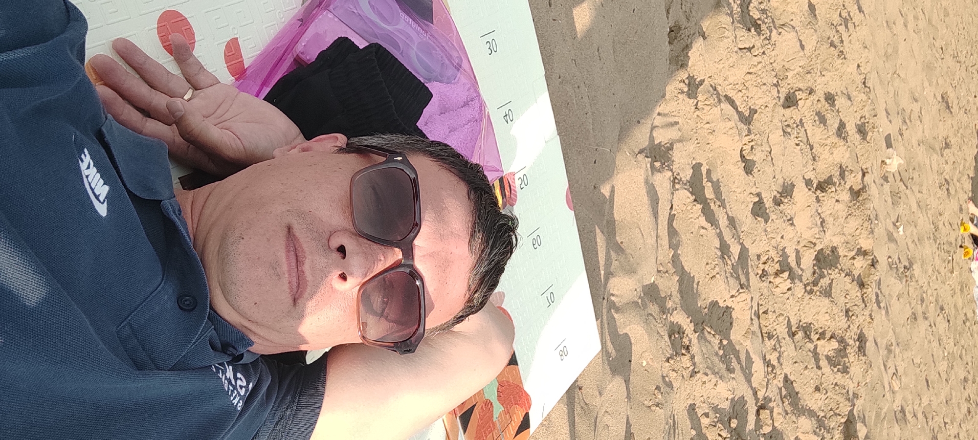 A man is lying on a mat on a sandy beach, possibly in Noviy, Russia. He is wearing a blue polo shirt with a Nike logo and sunglasses. His head is resting on his arm, and he appears relaxed. The mat has markings on it, possibly indicating measurements. In the background, there is sand and a hint of water, suggesting a beach setting. The lighting indicates it is daytime, likely sunny. The overall mood is one of leisure and relaxation.