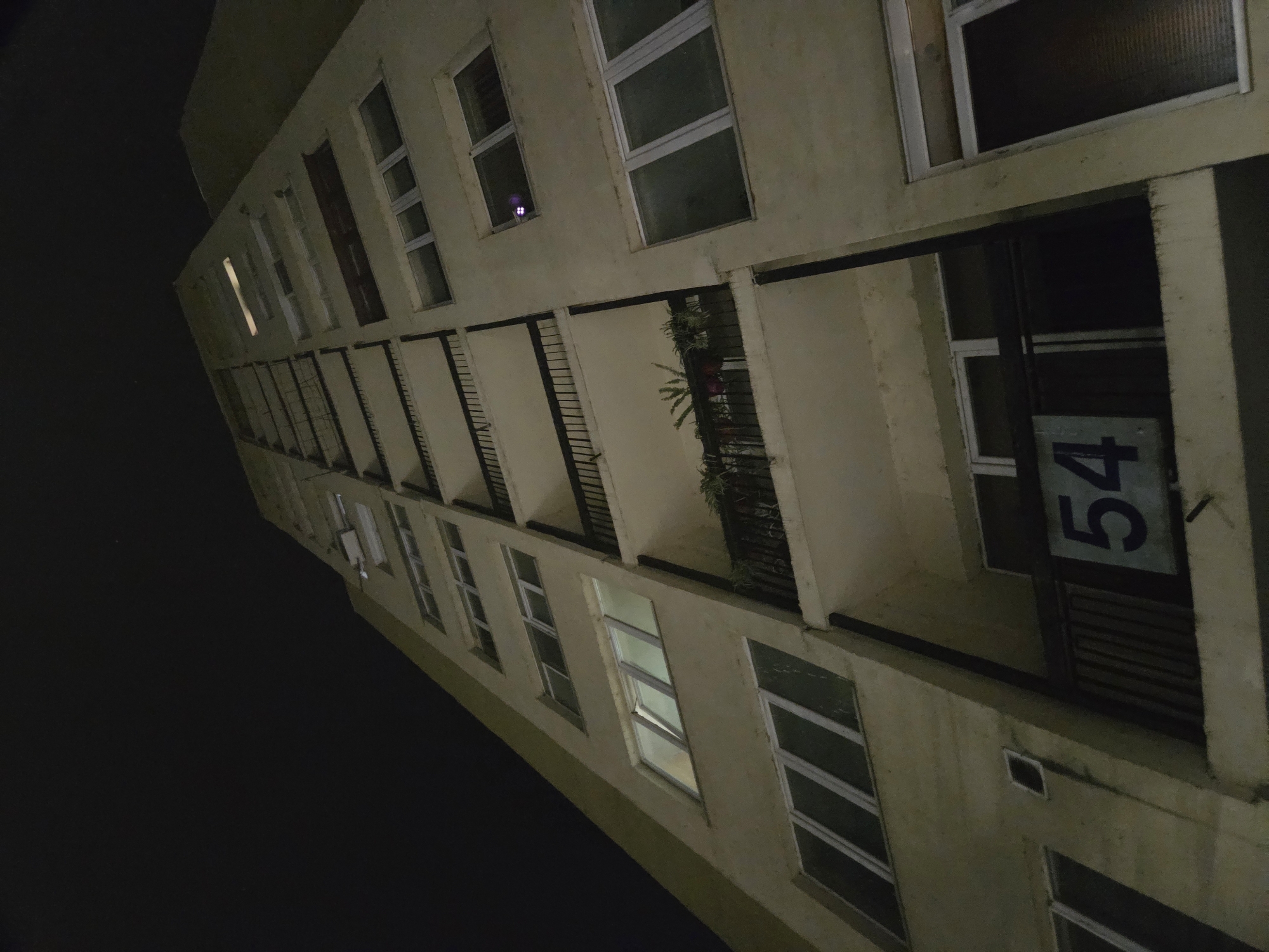 A low-angle, nighttime view of an apartment building is captured. The building is beige and appears to be multiple stories tall. Each level features rectangular windows and balconies, some of which have plants on them. The number "54" is prominently displayed on the side of one balcony. The sky is black, indicating it is night. The location is Pleven, Bulgaria.
