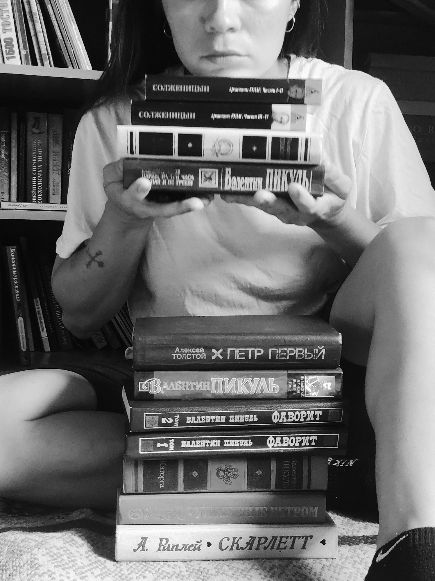 A black and white photograph captures a person, likely a woman, seated on a patterned surface in an indoor setting, surrounded by books. She holds a stack of four books to her chest, her gaze directed downwards, creating a contemplative or pensive mood. Her face is partially obscured by the books, but a slight furrow in her brow is visible. A tattoo of a cross is visible on her left forearm, and a hoop earring is in her right ear.
Another tall stack of books rests on the surface to her right. In the background, numerous books are arranged on shelves, suggesting a home library or reading space.
Visible text on the books she holds includes "СОЛЖЕНИЦЫН Архипелаг ГУЛАГ. Части I-II" (Solzhenitsyn, The Gulag Archipelago, Parts I-II) and "Части III-IV", as well as "Валентин ПИКУЛЬ" (Valentin Pikul). The stack on the floor features "Алексей ТОЛСТОЙ X ПЕТР ПЕРВЫЙ" (Alexei Tolstoy, Peter the Great), several more by "ВАЛЕНТИН ПИКУЛЬ" including "НА ЗАДВОРКАХ ВЕЛИКОЙ ИМПЕРИИ" (In the Backyards of a Great Empire), "ФАВОРИТ" (The Favorite) volumes 1 and 2, and "Каторга". The bottom book is "А. Риплей • СКАРЛЕТТ" (A. Ripley • Scarlett). Bookshelves in the background display titles like "1500 ТОСТО", "Комнатные растения" (Indoor plants), "ОБЩЕСТВОЗНАНИЕ" (Social Studies), and "НОВЕЙШИЙ СПРАВОЧНИК НЕОБХОДИМЫХ ЗНАНИЙ" (The Newest Reference Book of Essential Knowledge).