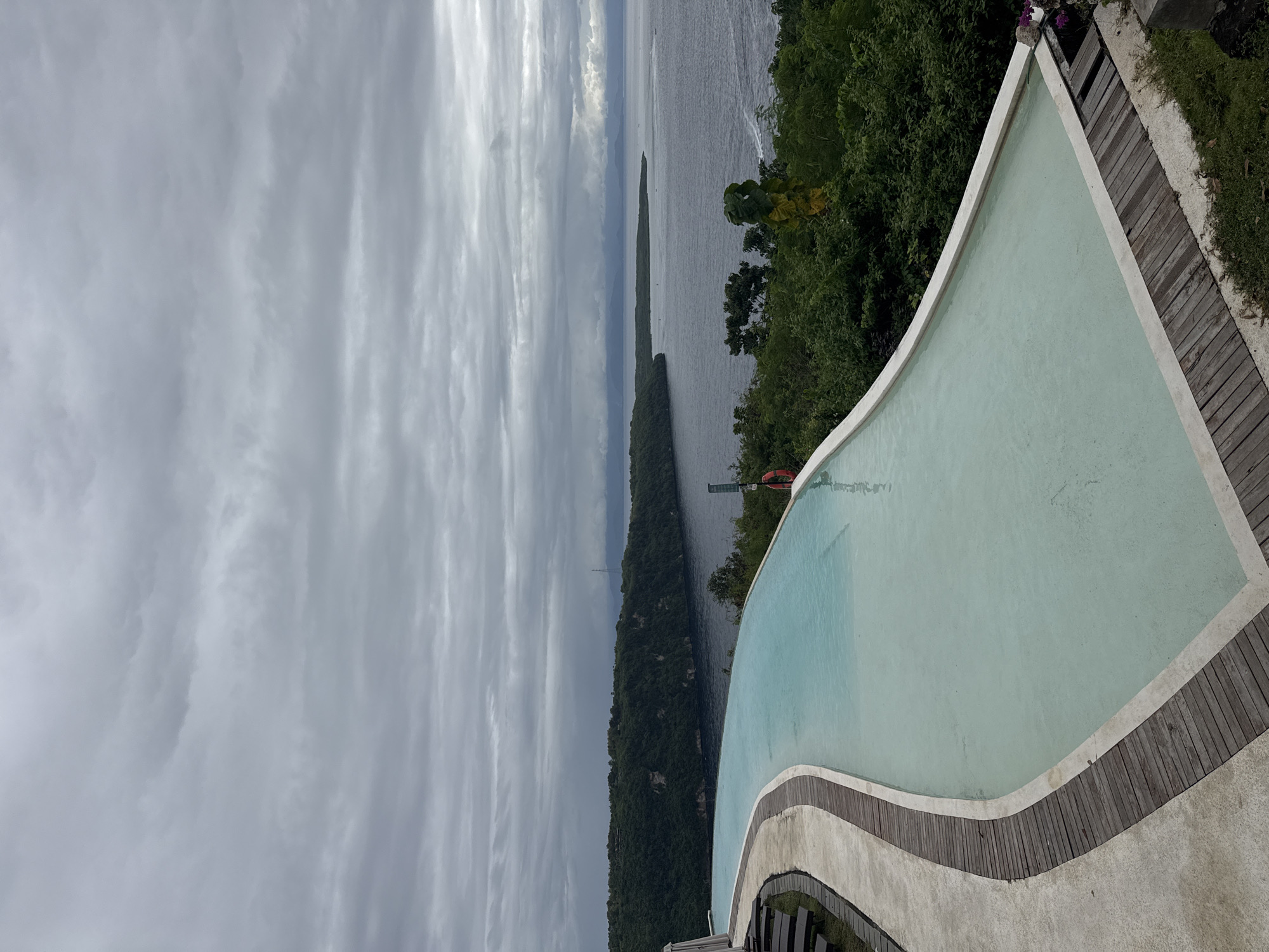 The image presents a view of a rectangular pool with rounded edges, adjacent to wooden decking and lush greenery. In the background, a long, thin landmass stretches into a body of water under a cloudy sky. The location is Nusa Penida, Indonesia, inferred from the geographical features visible in the image. A red life preserver is attached to a pole near the pool.