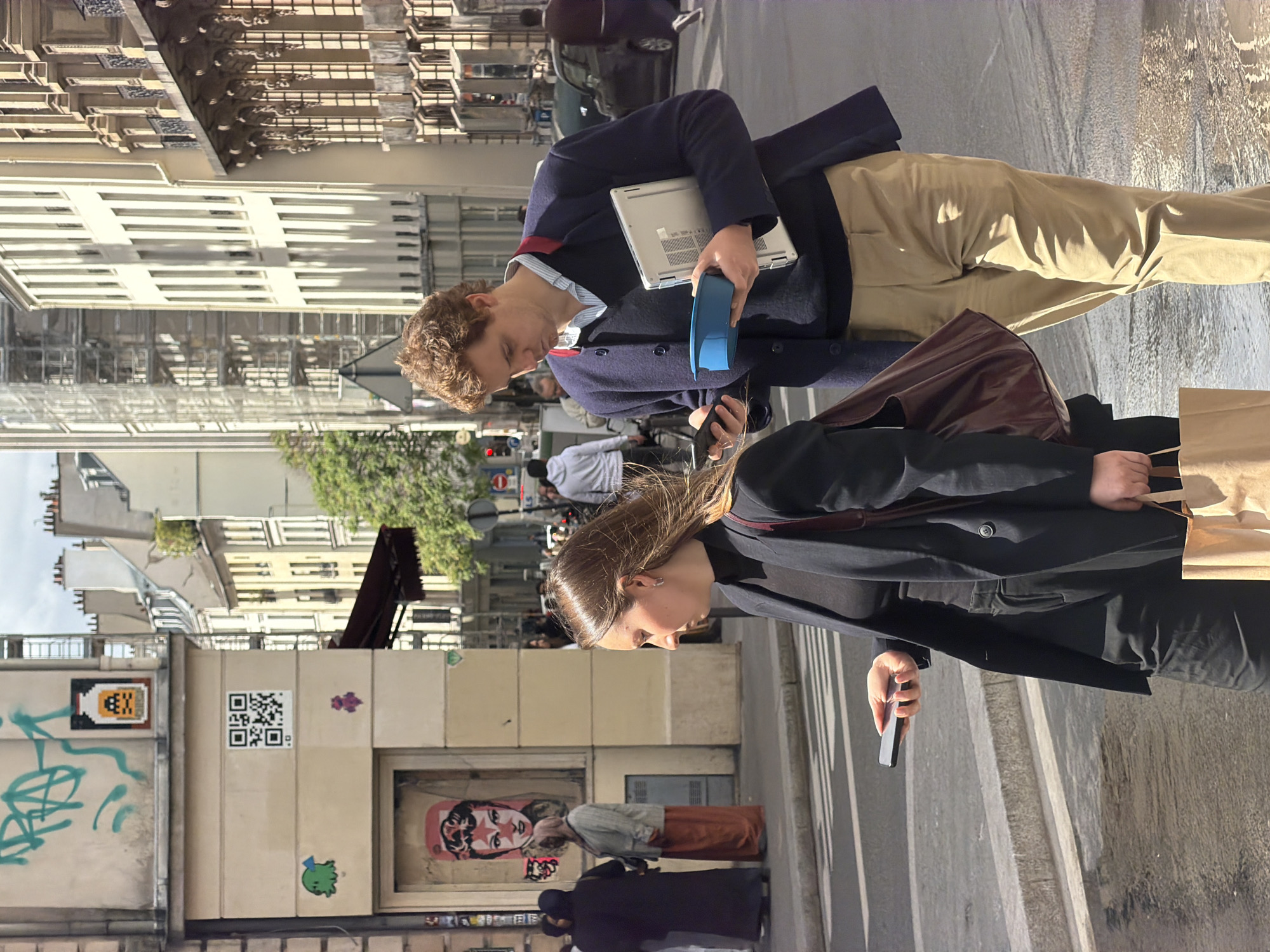 The image depicts a daytime street scene in what appears to be an urban setting like Paris, given the architectural style of the buildings. Two main individuals are walking in the foreground on a paved sidewalk. The person on the left, a man with curly brown hair, wears a navy blue jacket over a striped shirt and khaki pants. He is holding a silver laptop in his right arm and a blue cylindrical object in his left hand, while also looking down at a smartphone. The person on the right, a woman with long brown hair, wears a black coat and carries a dark brown shoulder bag, as well as a light brown paper bag. She is also engrossed in her smartphone, held in her right hand.
The background features classic, multi-story buildings with numerous windows and some balconies, indicative of Parisian architecture. On a wall to the left of the subjects, various pieces of street art are visible. These include a pixelated yellow character resembling a Space Invader, a black and white QR code, a small purple design, green graffiti tags, and a prominent framed artwork of a stylized face, possibly a woman, with a star on her forehead. Another small green monster-like character is also on the wall. Several other pedestrians are scattered in the midground and background. The street to the right appears wet or reflective, possibly from recent rain or dampness. The overall lighting suggests a bright, sunny day. No explicit text is readable from the QR code or the graffiti.