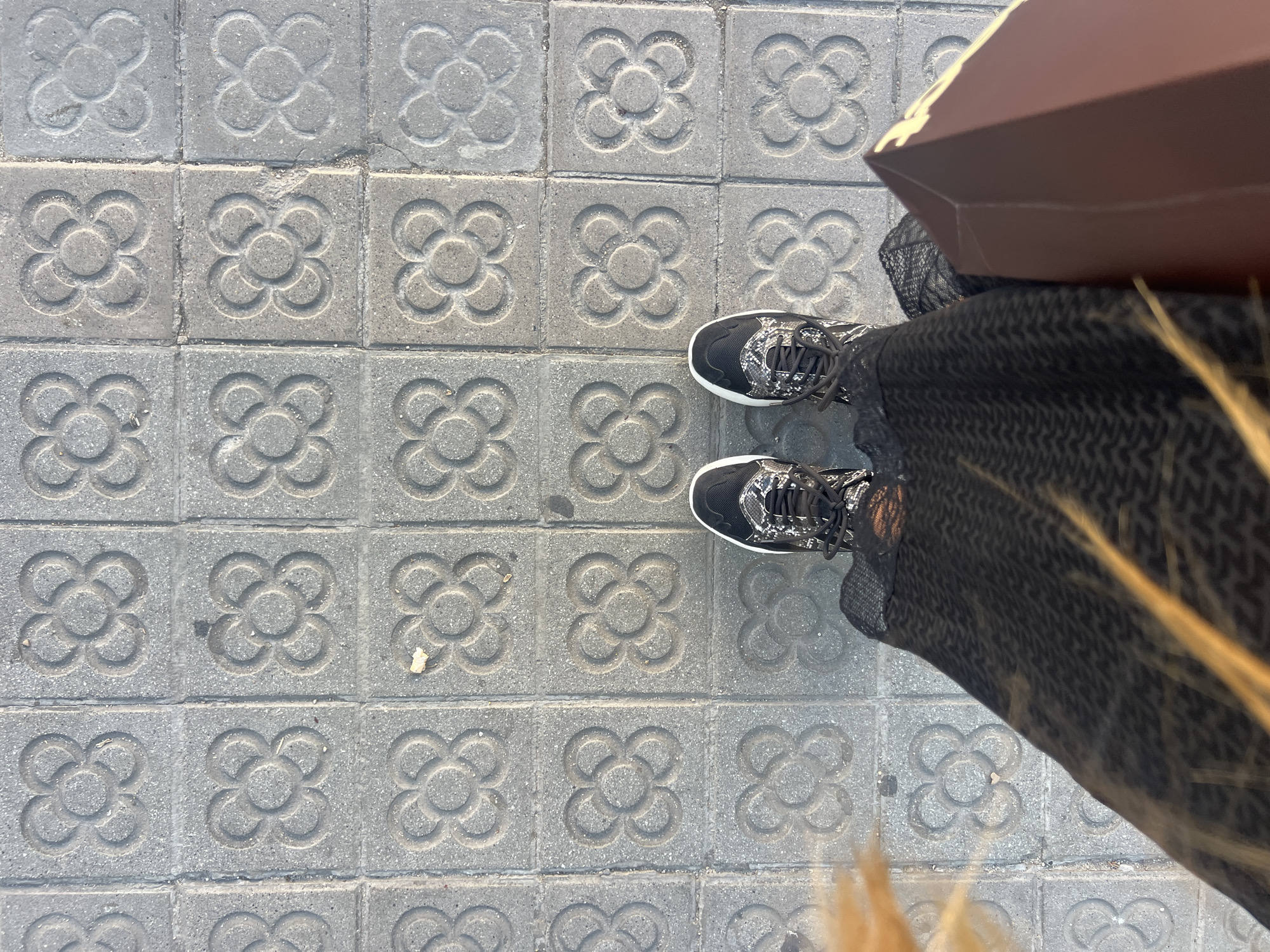 An overhead view captures the lower torso and feet of a person standing on a distinctively patterned sidewalk in Barcelona, Spain. The gray concrete sidewalk is paved with square tiles featuring the embossed "Flor de Barcelona" (Flower of Barcelona) design, a four-petal floral motif with a central circle, characteristic of the city's urban landscape. The tiles show varying degrees of wear and minor debris.
The person wears black sneakers with white sole trim, a black and white snakeskin-like pattern on the upper, and black laces. A dark, possibly black or very dark gray, textured fabric from a skirt or dress is visible over the legs. Blondish or light brown hair cascades along the left edge of the frame. On the right side, a rectangular brown box or bag is held, partially obscuring the right foot. The scene is illuminated by bright, natural light, casting subtle shadows and emphasizing the pavement's texture.