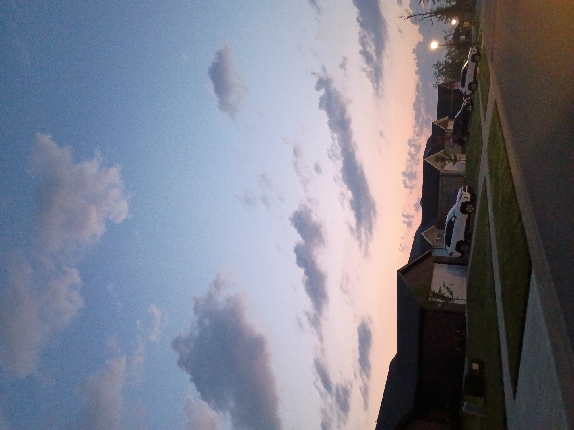 The image depicts a suburban street scene at dusk, with a row of houses lining the right side of the frame. The sky is a gradient of blue, transitioning to pale pink and orange near the horizon, with scattered clouds. Several vehicles are parked in driveways or on the street. The foreground shows a paved road and a grassy area. This scene is located in the city of Richmond, United States.