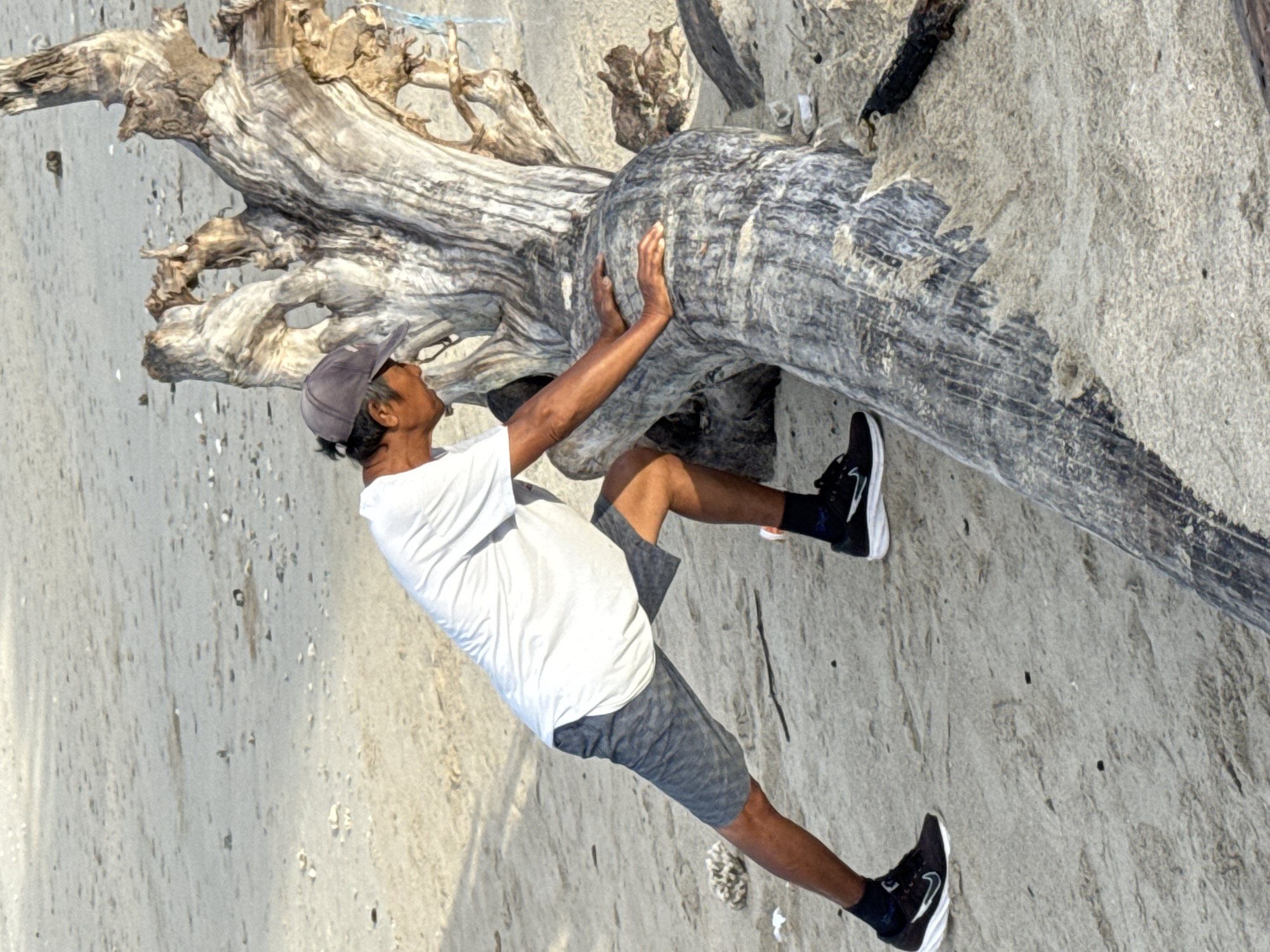 A man wearing a white T-shirt, gray shorts, a gray cap, and black athletic shoes is standing on a sandy beach next to a large piece of driftwood. He is touching the driftwood with his hands and looking up at it. The background is a sandy beach with the ocean in the distance. The image was taken in Denpasar, Indonesia.